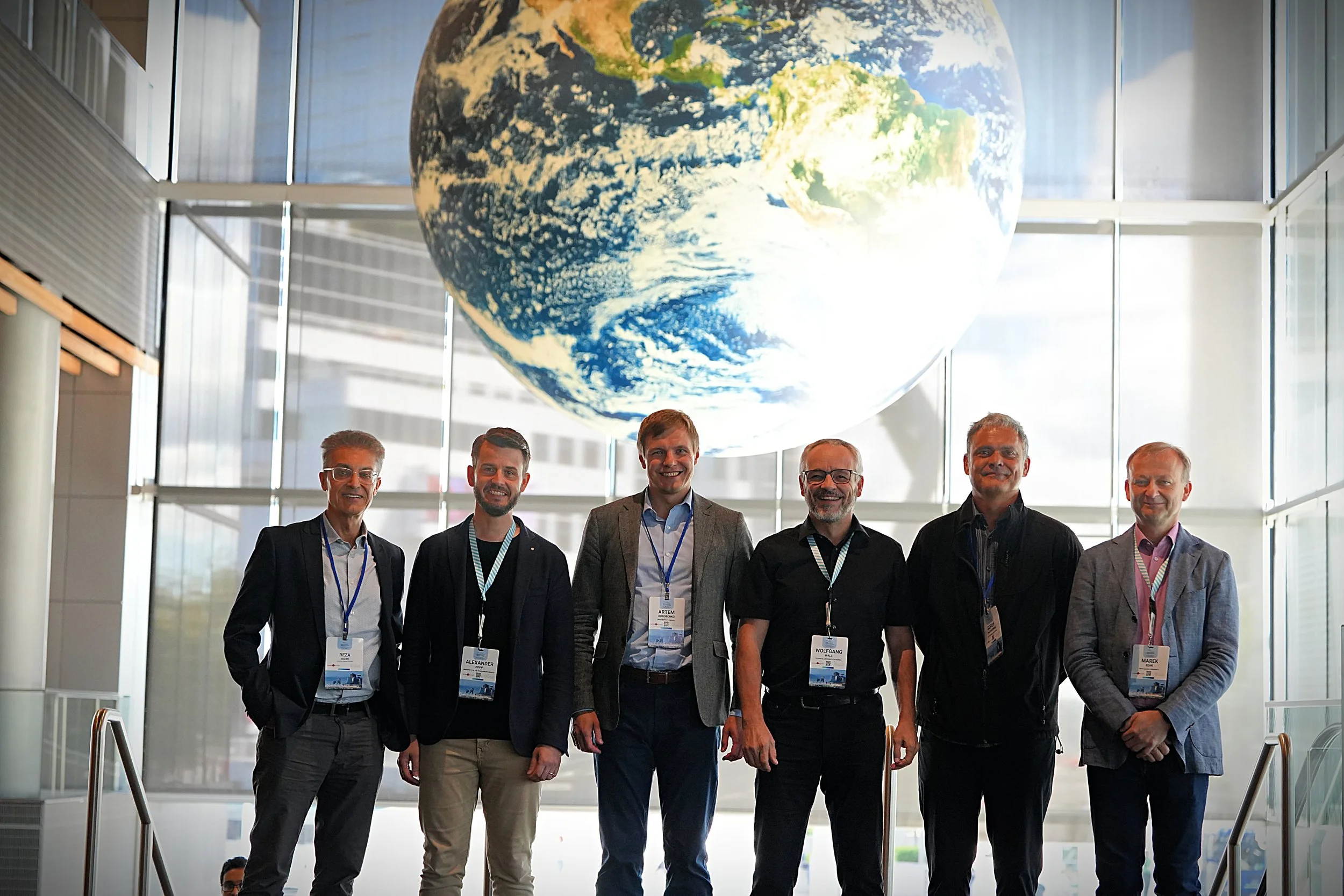 Group of six men standing inside a building with a large globe of the Earth hanging from the ceiling behind them.