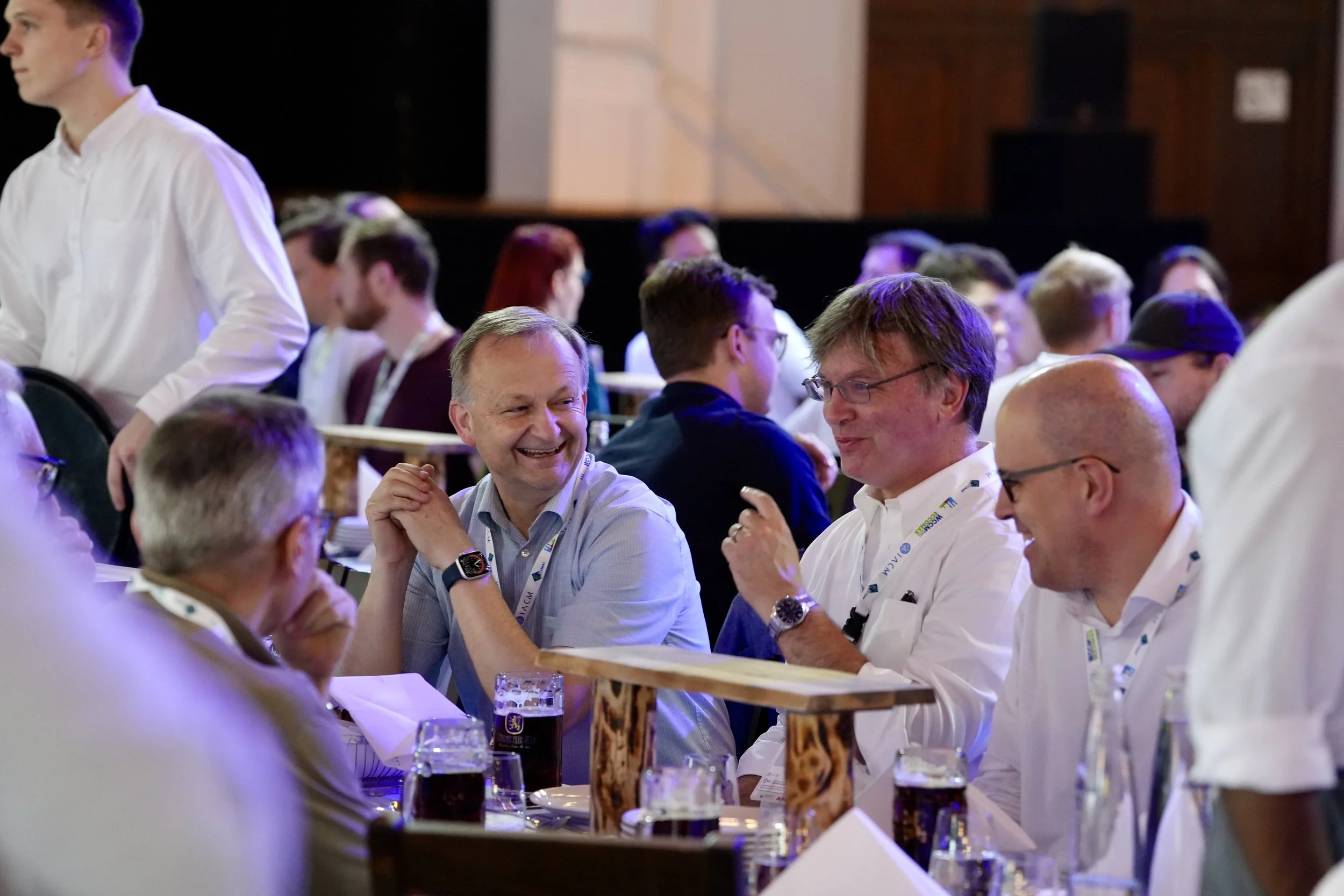 People sitting at a long wooden table engaging in conversation, with drinks and glasses in front of them, at a social or professional event.