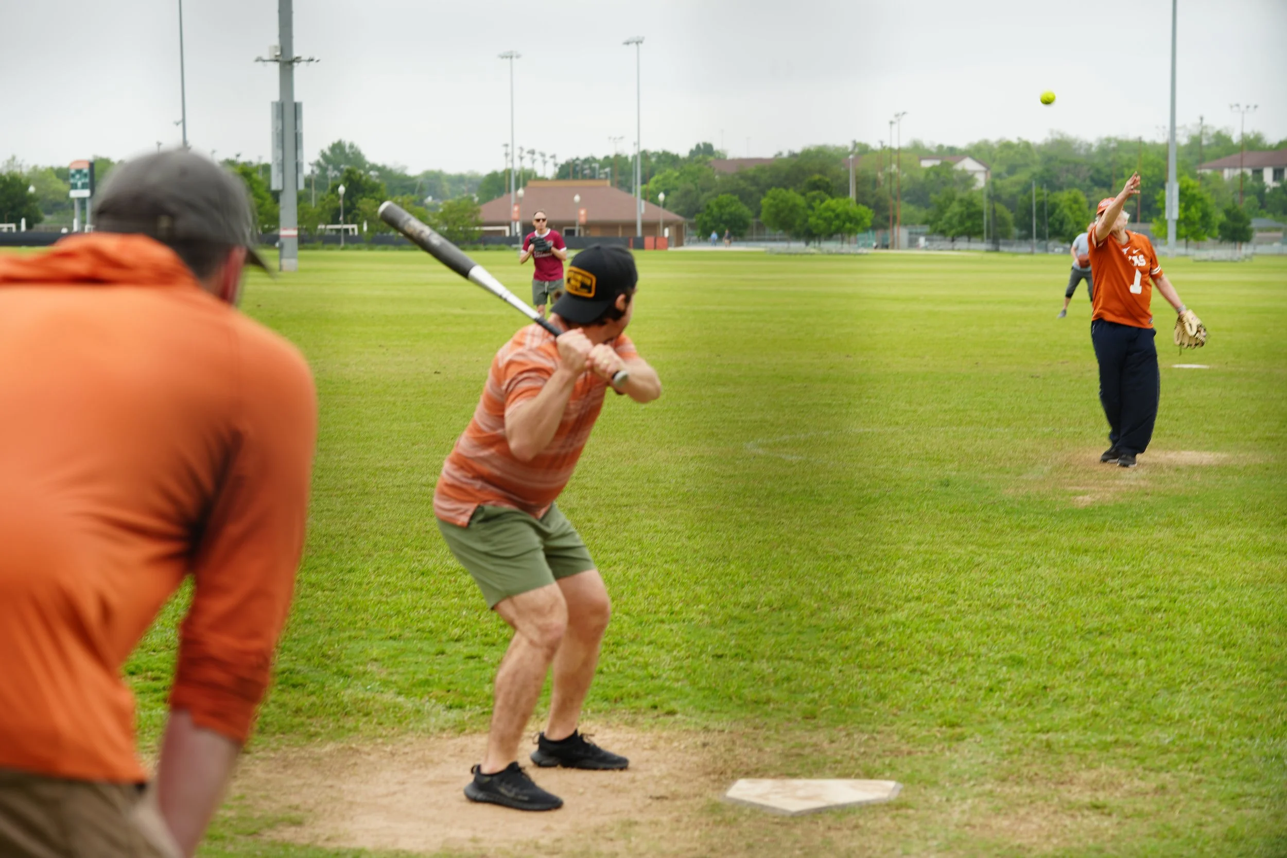 A group of people playing baseball on a grassy field, with one person at bat, others watching, and a player throwing the ball.