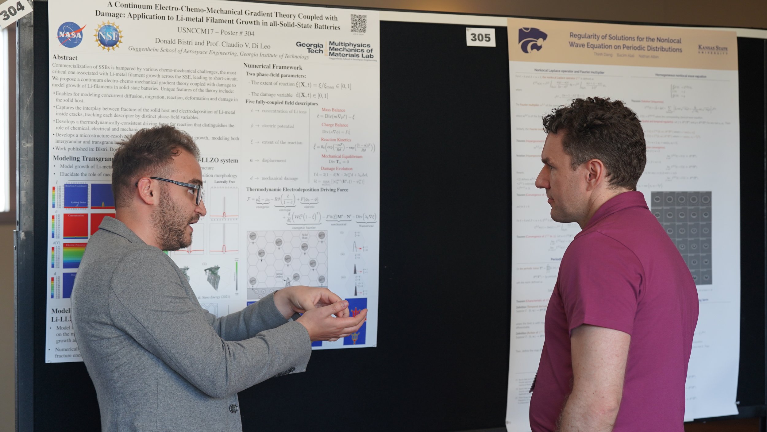 Two men are standing in front of research posters at a conference, with one presenting and the other listening. The posters contain scientific equations and diagrams.