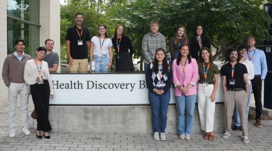 Team of research associates standing and sitting outside in front of a sign that reads 'Health Discovery' on a concrete wall, with trees and a building in the background.