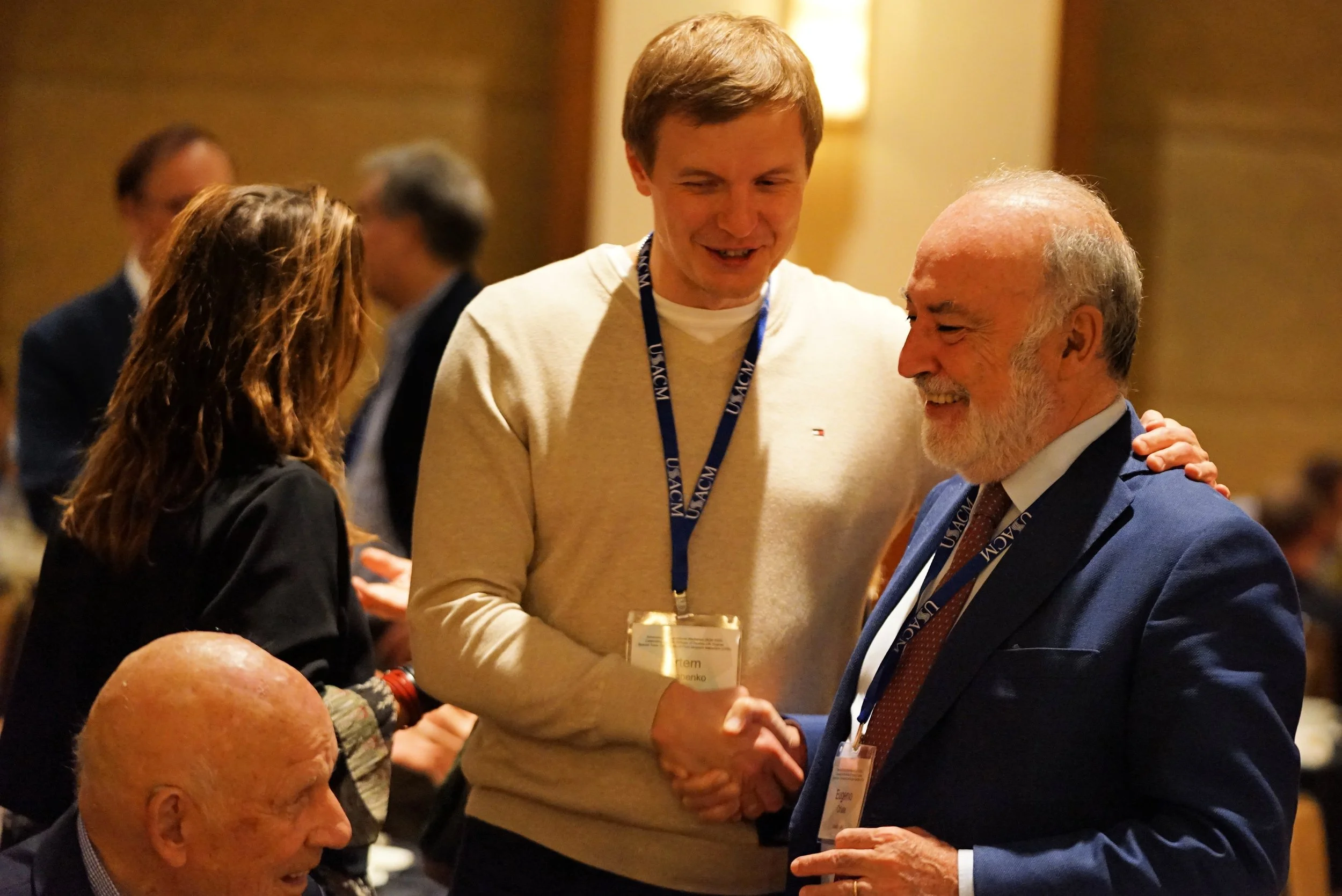 Two men shaking hands at a conference or event, with a woman nearby and other attendees in the background.