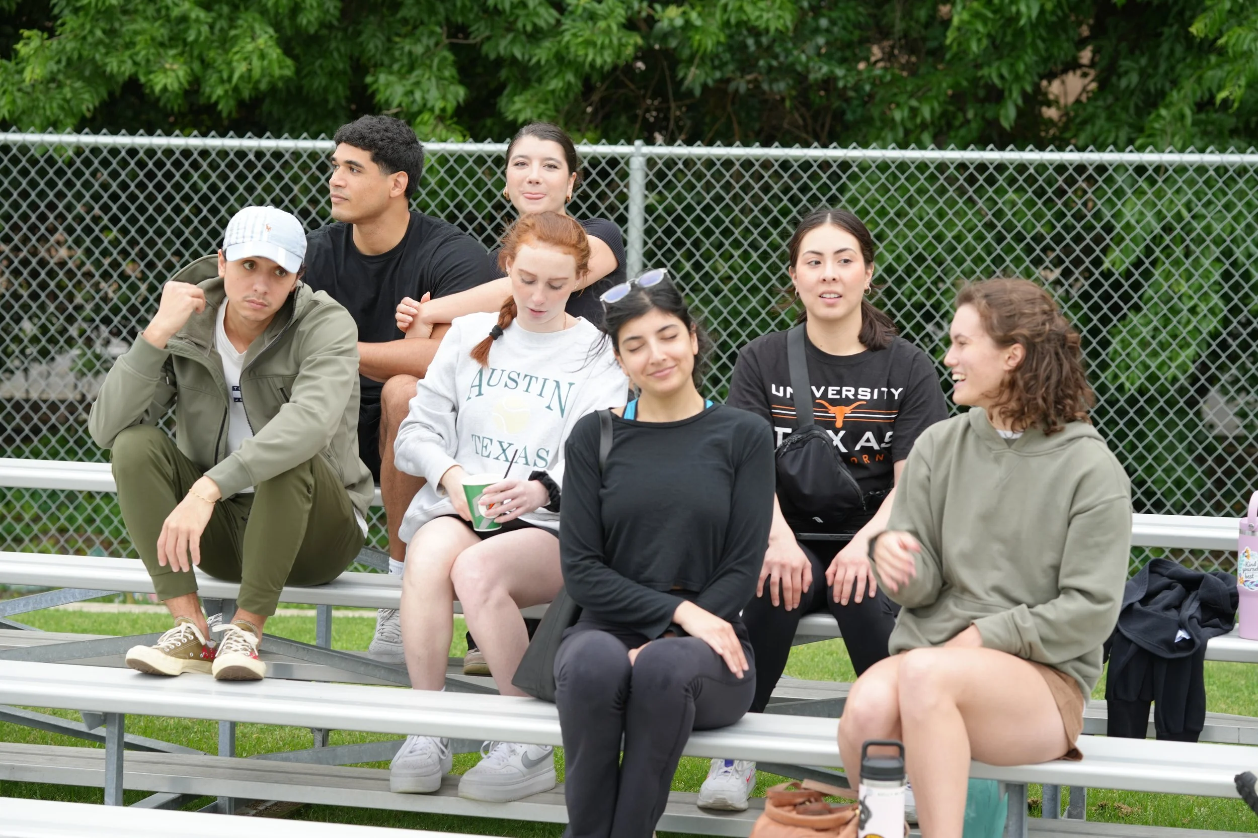 Group of seven young people sitting on bleachers outdoors near a chain-link fence, with trees behind them. Some are talking, others are looking around. They appear relaxed and casual, dressed in T-shirts, hoodies, and casual pants.