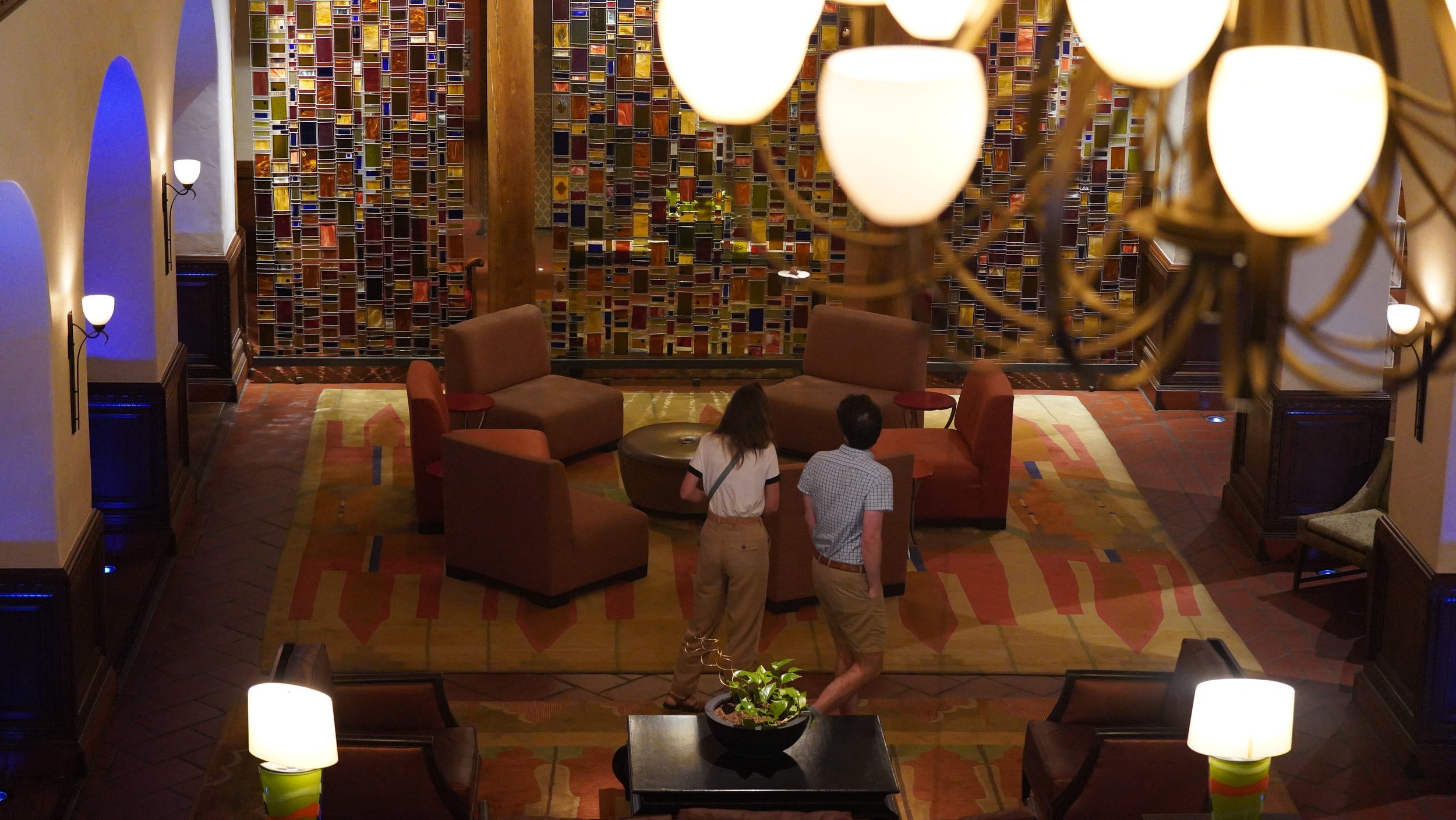 Interior of a warmly lit hotel lobby with a colorful mosaic wall, chairs, tables, and a chandelier. Two people are standing near the center, engaged in conversation, with other furniture and decor visible.