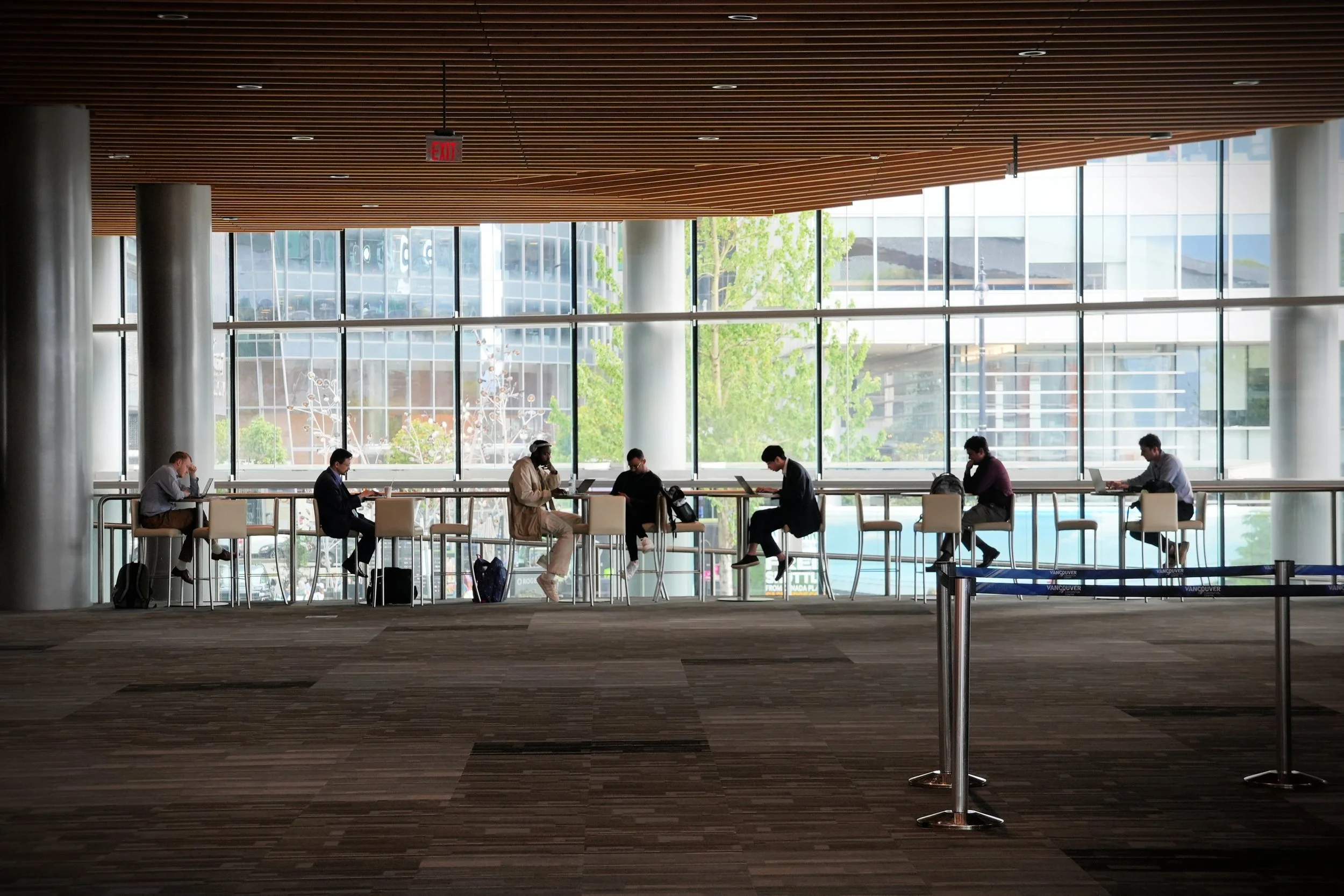 Interior of a modern building with floor-to-ceiling windows, showing people sitting on chairs, some working on laptops, with a cityscape and greenery outside.