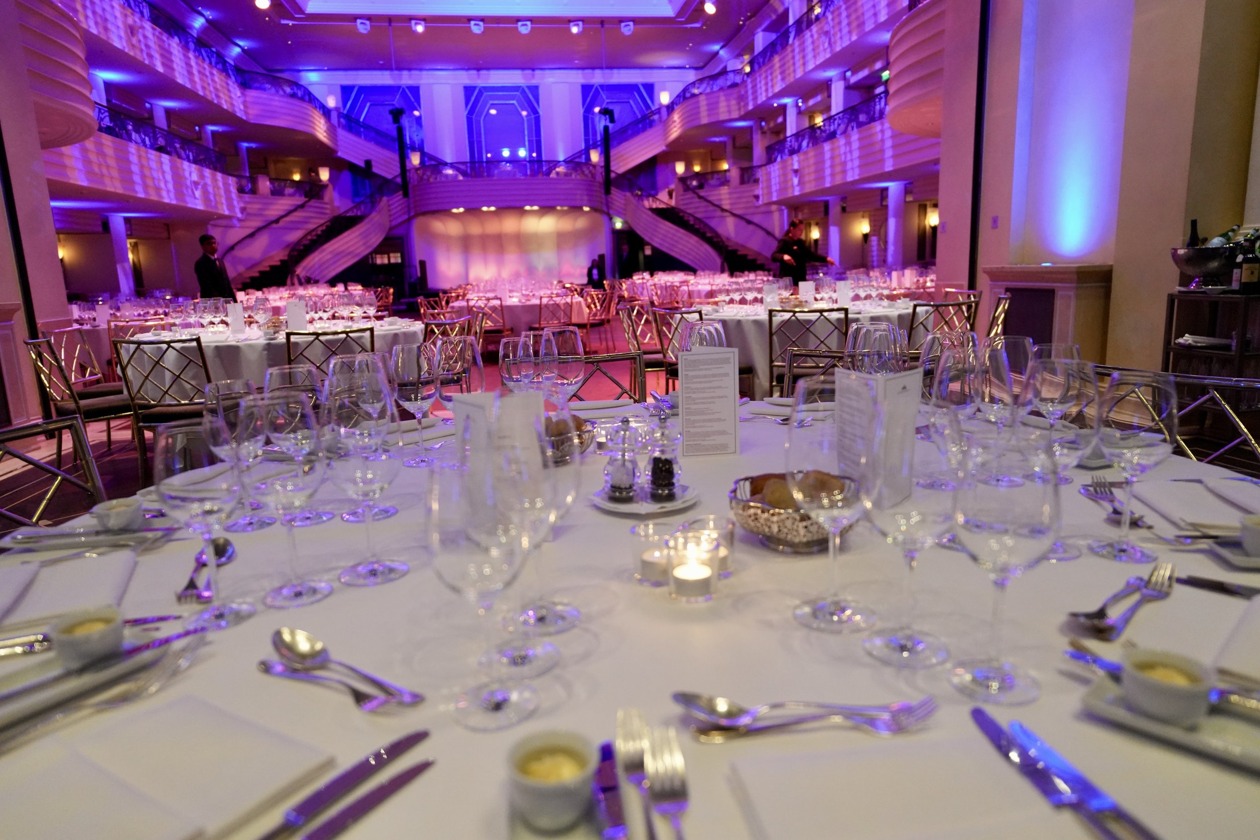 Elegant banquet hall with round tables covered in white tablecloths, set with wine glasses, plates, silverware, and candles. The room has a grand staircase, purple and blue lighting, and a stage at the back.