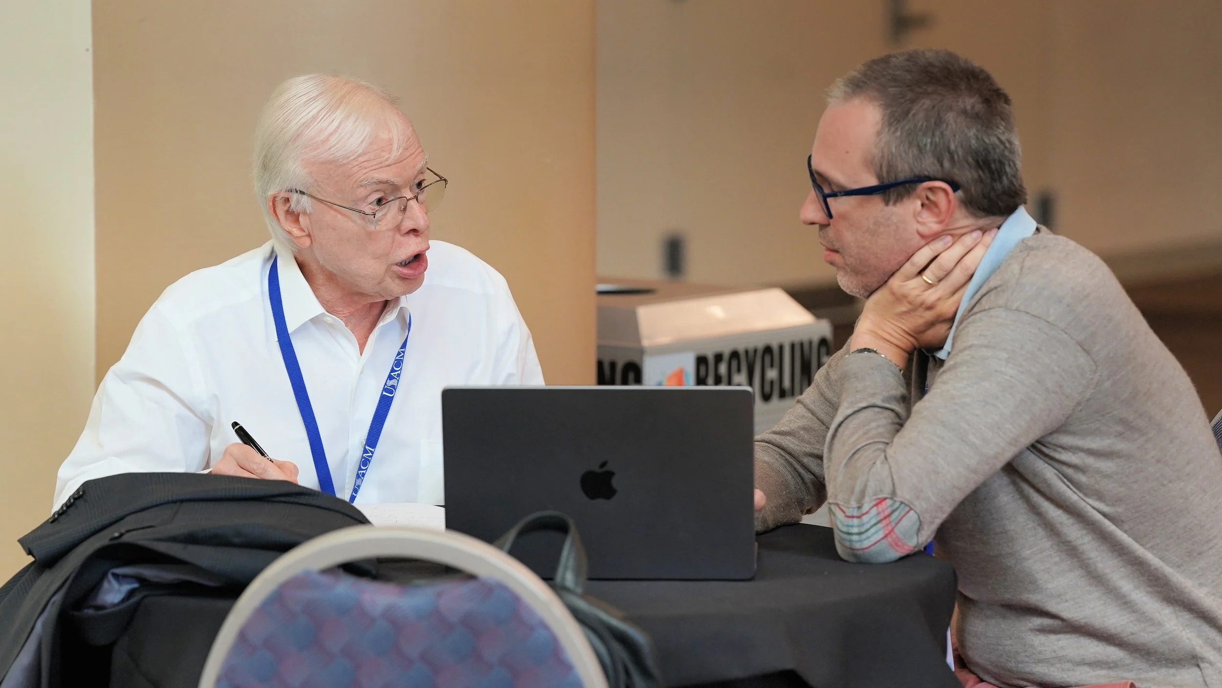 An elderly man with white hair and glasses wearing a white shirt and a blue lanyard, speaking to a middle-aged man with glasses and brown hair, who is resting his neck on his hand, at a table with a laptop and a recycling bin in the background.