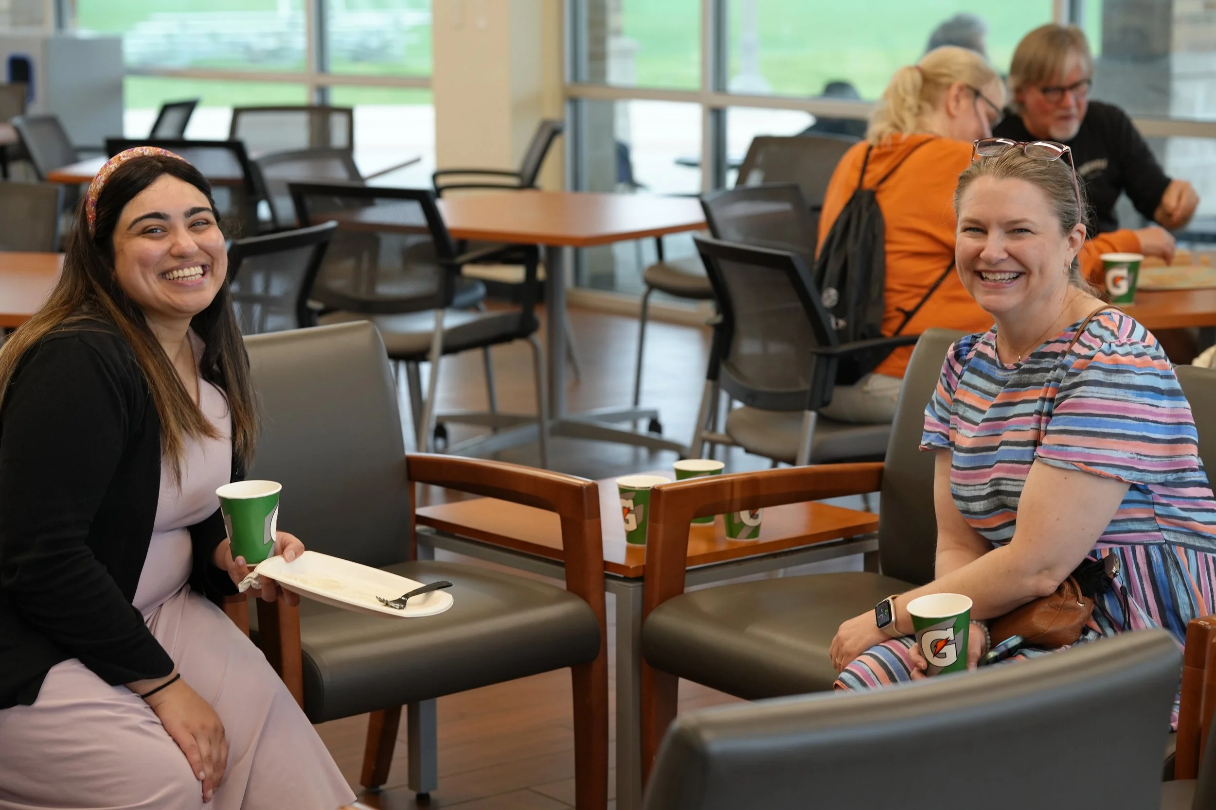 Four women sitting and laughing in a casual meeting room, some holding Cups of Gatorade, with large windows in the background.