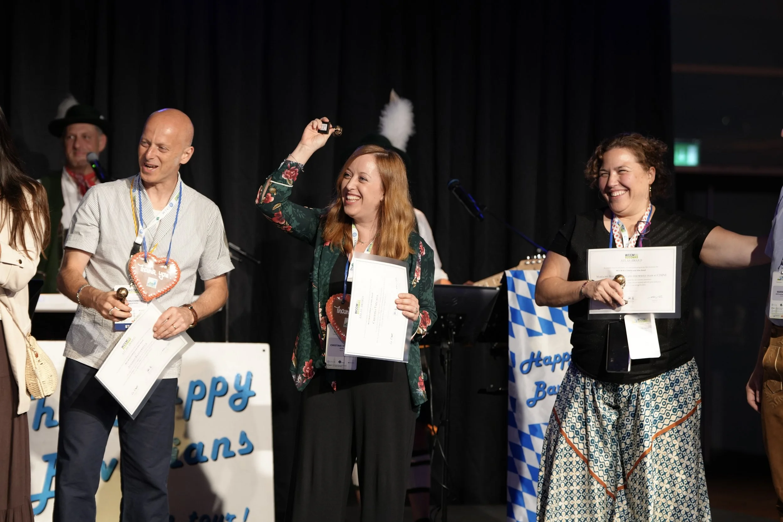 Several people on stage celebrating, holding certificates and flowers, with a black curtain background and decorations including a blue and white checkered cloth and a sign that reads 'Happy'.