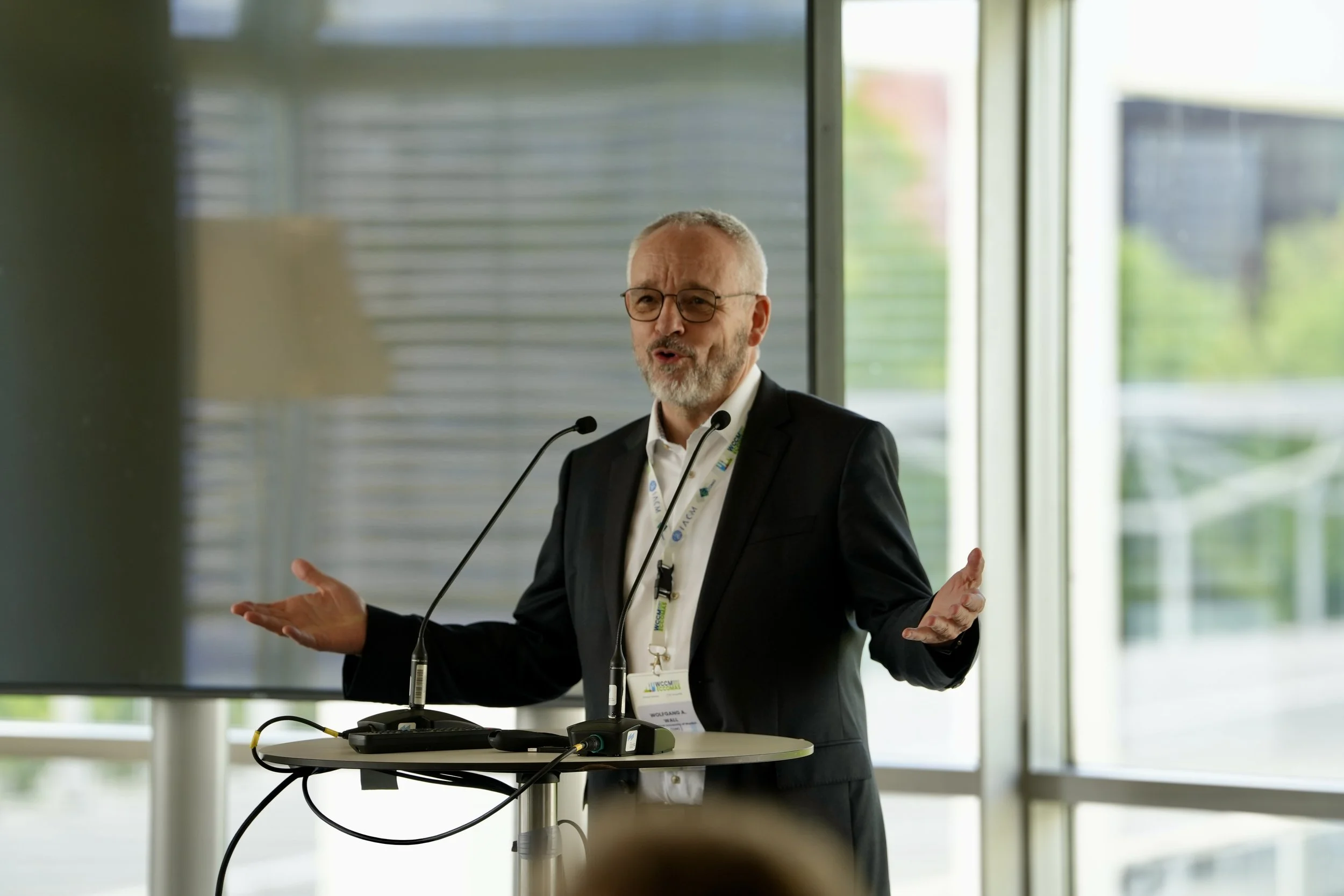 A middle-aged man with glasses and a beard, dressed in a dark suit and white shirt, giving a presentation at a conference. He is standing behind a small round table with microphones and a lapel mic. The background features large windows with a modern building and greenery outside.