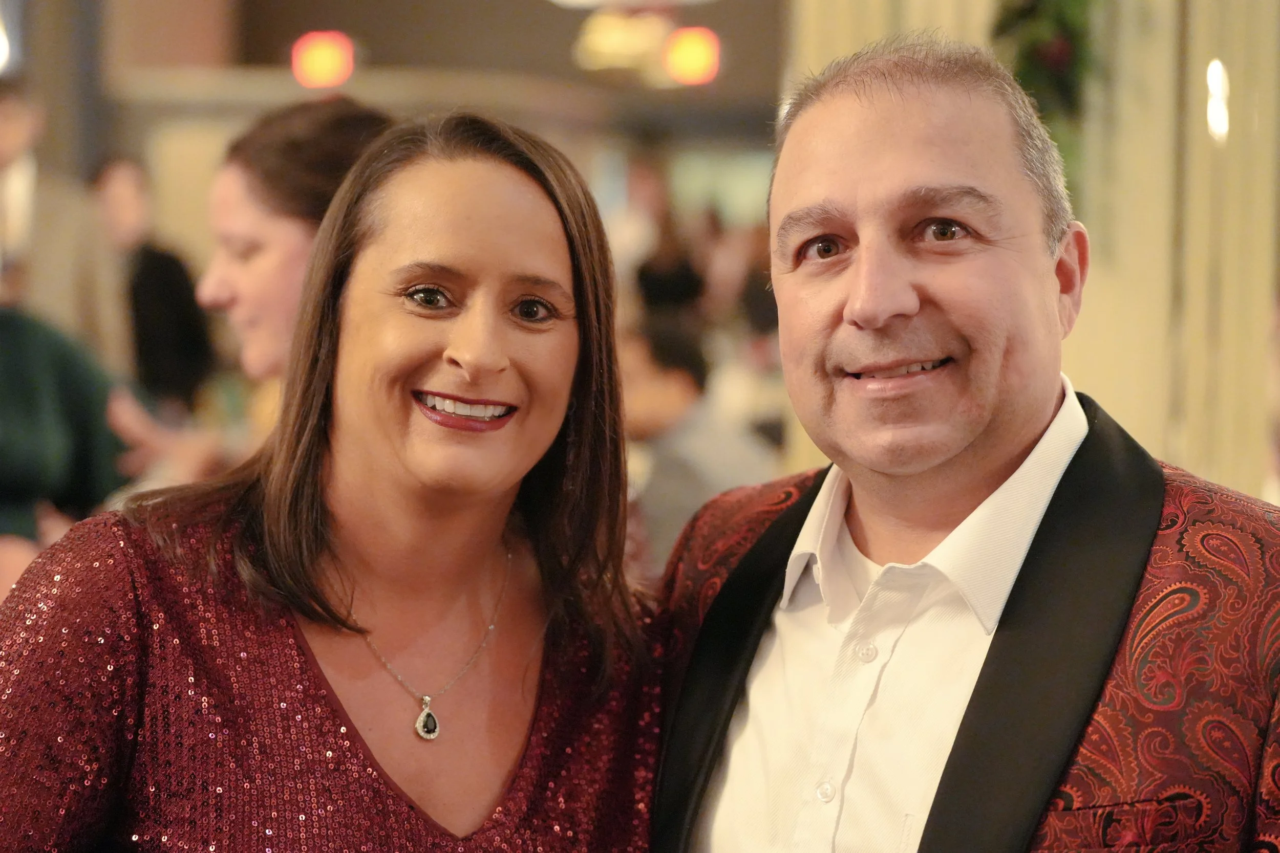 A woman with long brown hair, wearing a red sequined dress and a silver necklace with a pearl pendant, standing next to a man with short light-colored hair, wearing a tuxedo with a red and black patterned shawl collar, at an indoor event.