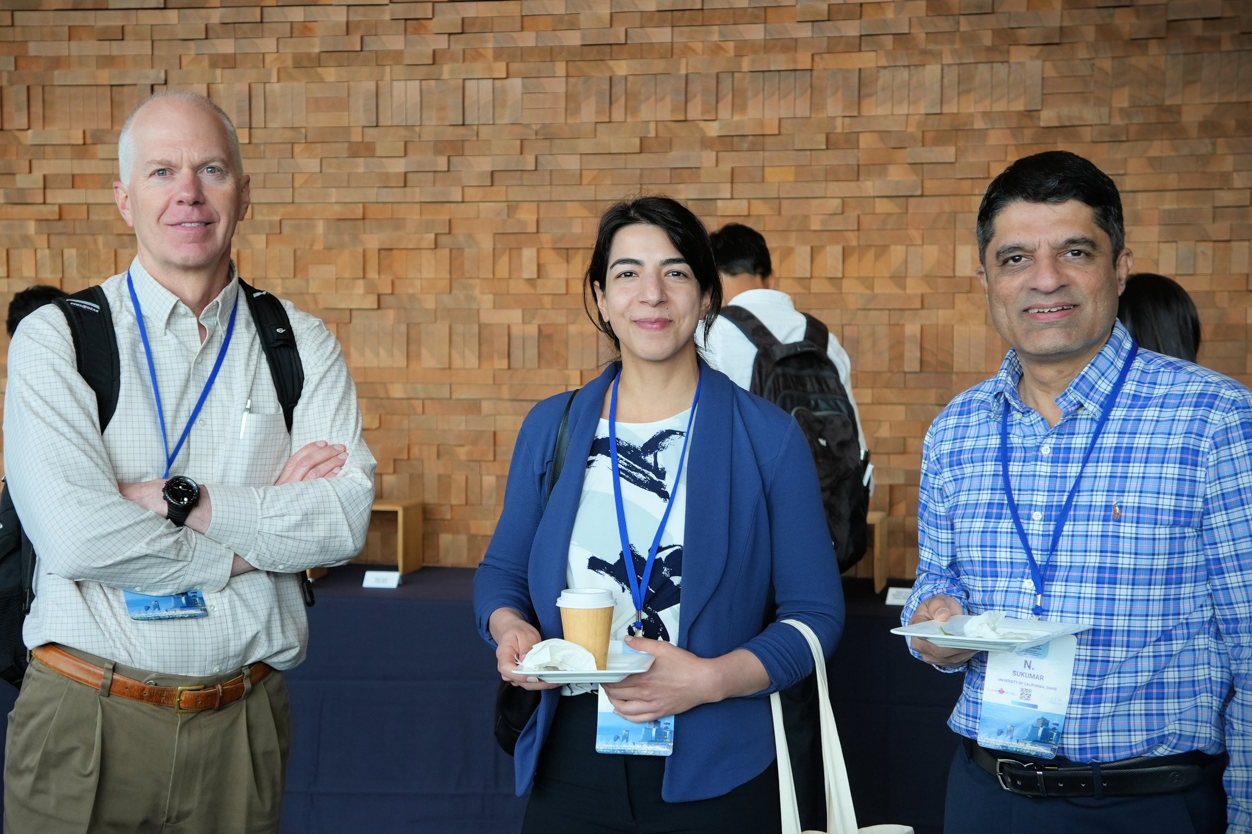 Three people standing in front of a brick wall, smiling at the camera. The person on the left is a man with short, light-colored hair, wearing a light-colored shirt, carrying a backpack, and with crossed arms. The person in the middle is a woman with dark hair, wearing a blue jacket and holding a coffee cup and a plate with food. The person on the right is a man with dark hair, wearing a blue checkered shirt, holding a plate with food.