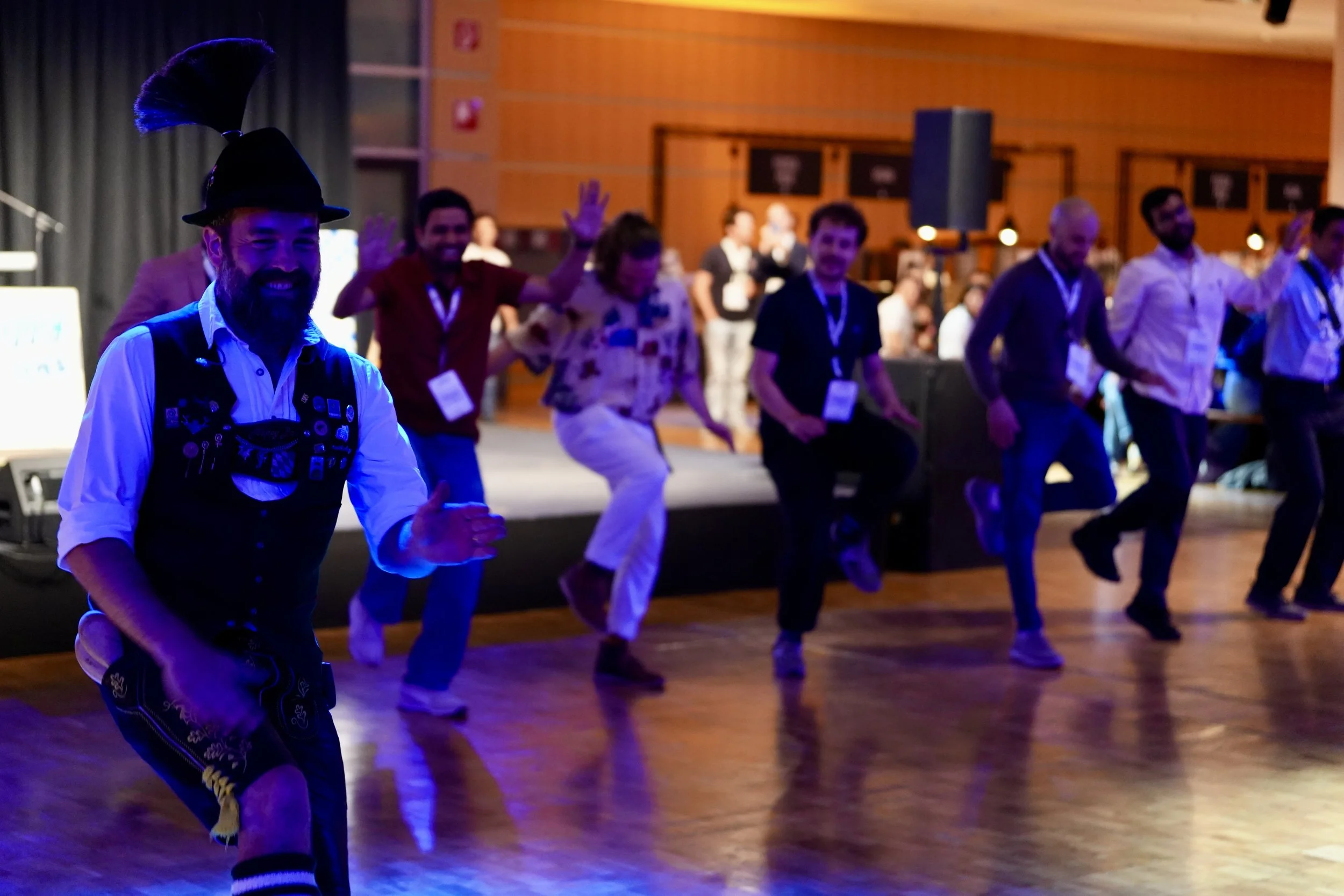 Group of people dancing in a dimly lit room, some smiling and waving their arms, with a man in the foreground wearing a black hat with a feather and decorated vest. They are at a conference dinner.