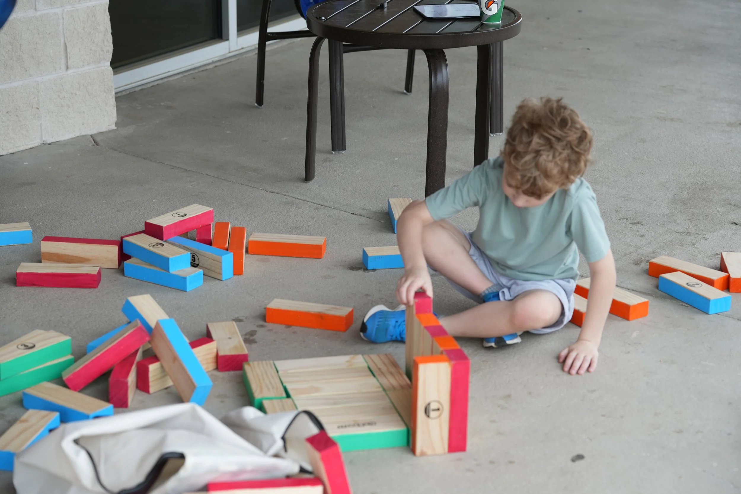 Child with curly hair sitting on the sidewalk playing with colorful wooden blocks, some stacked and some scattered around, beside a black patio table with a Gatorade bottle and a snack on it.