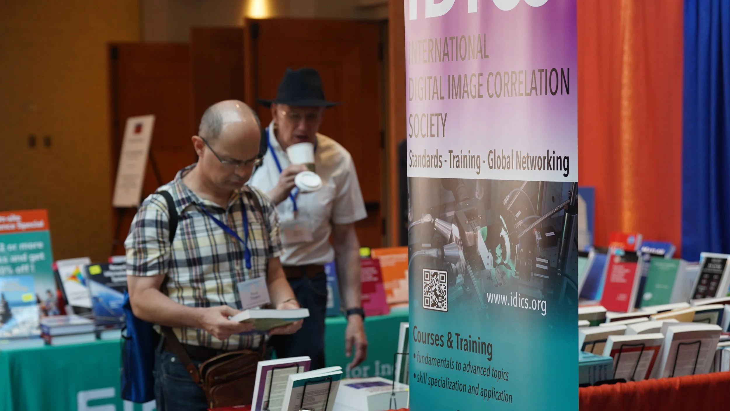 Two men at an international digital image correlation conference booth. One man is reading a booklet, the other is drinking from a cup. The booth features a tall, colorful banner with text about digital image correlation society and a QR code. The background shows a table with books and other materials.