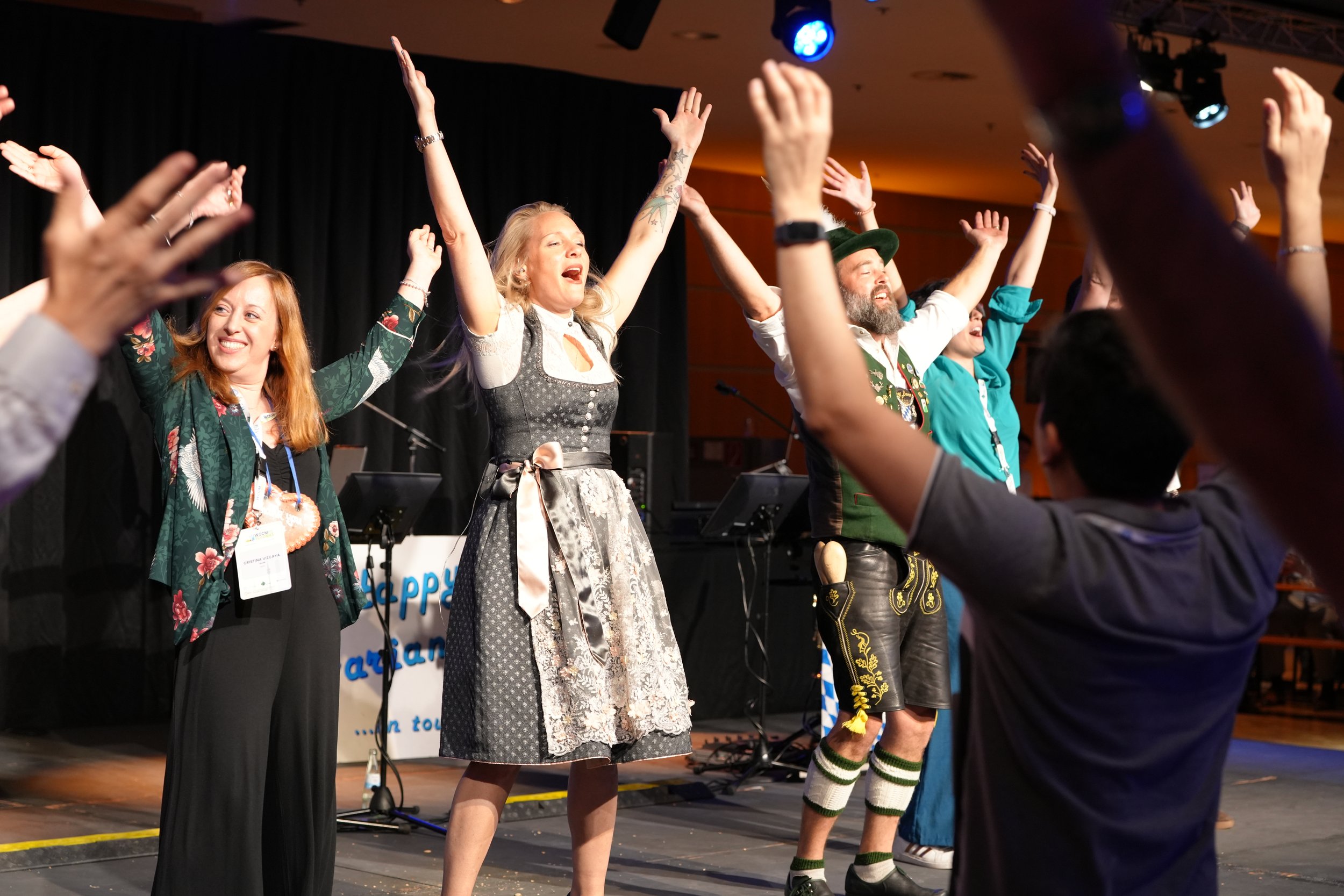 People on stage celebrating with their hands raised, some in traditional German attire, with a black curtain and stage lighting in the background.