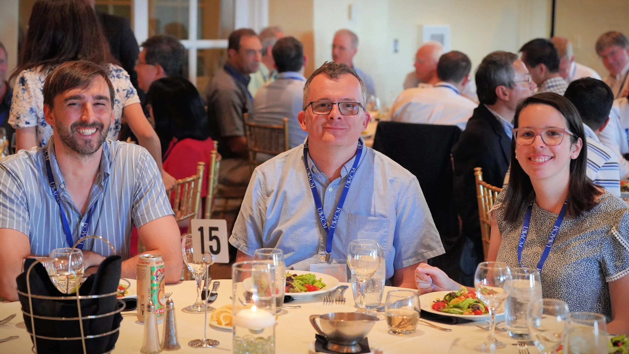 Three people sitting at a round banquet table during a formal event, smiling at the camera. The table is set with salad, glasses, and silverware, and has a table number 15. People in the background are engaged in conversation.