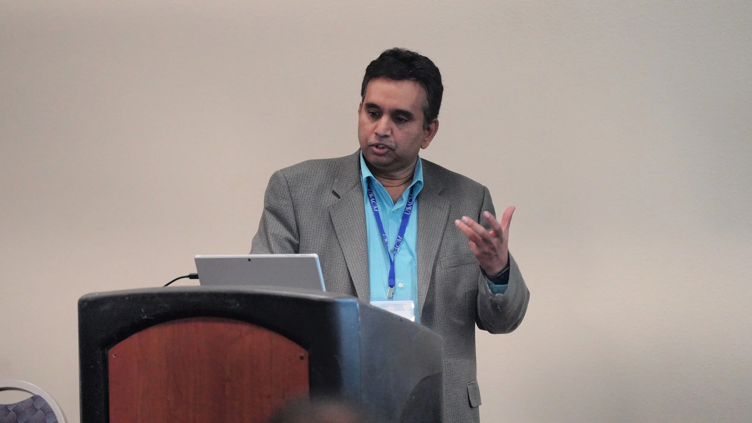 A man in a gray suit and blue shirt speaking during a presentation, standing behind a laptop and gesturing with his right hand in front of a blank wall.