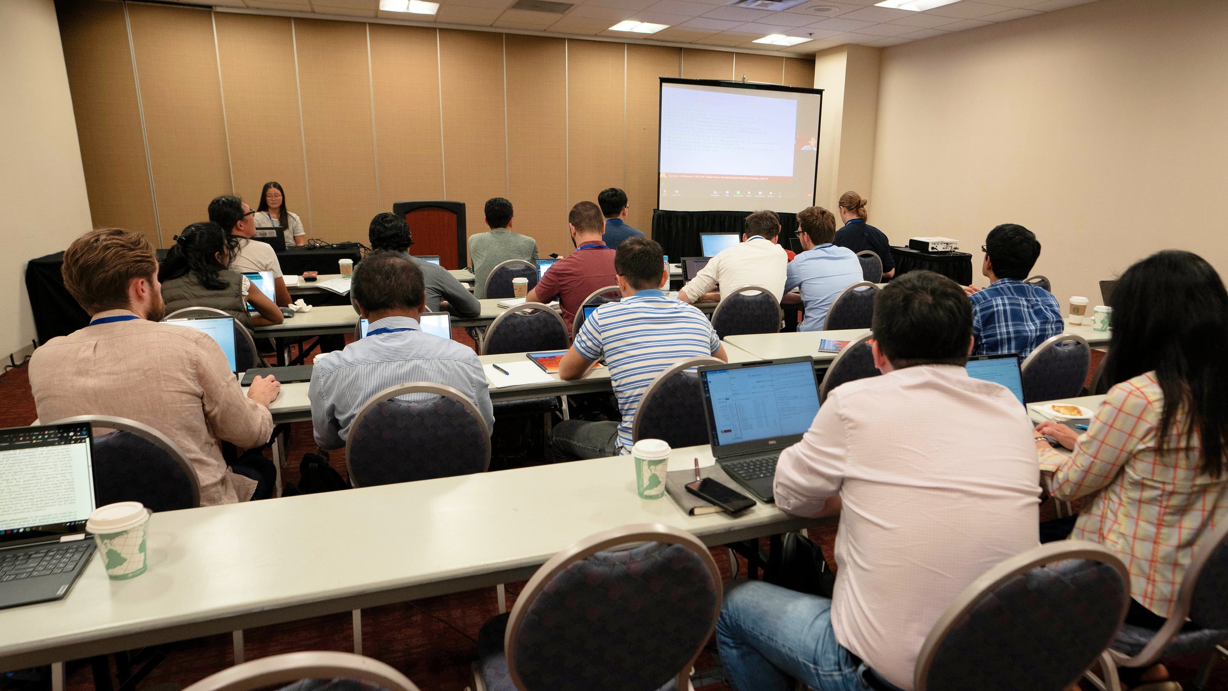 A conference room filled with attendees sitting at tables, facing a large screen at the front where a presentation is being projected. The attendees are using laptops and notebooks, with coffee cups on the tables.