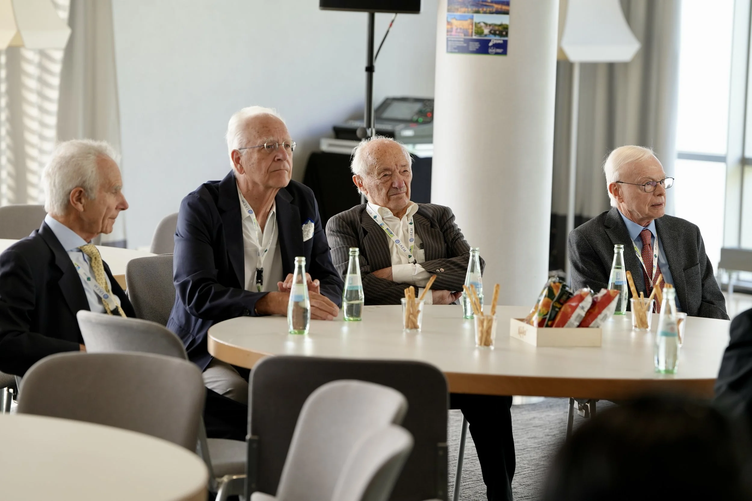 Four elderly men sitting at a conference table in a modern, well-lit room, wearing suits and ID badges, with beverages and snacks on the table.