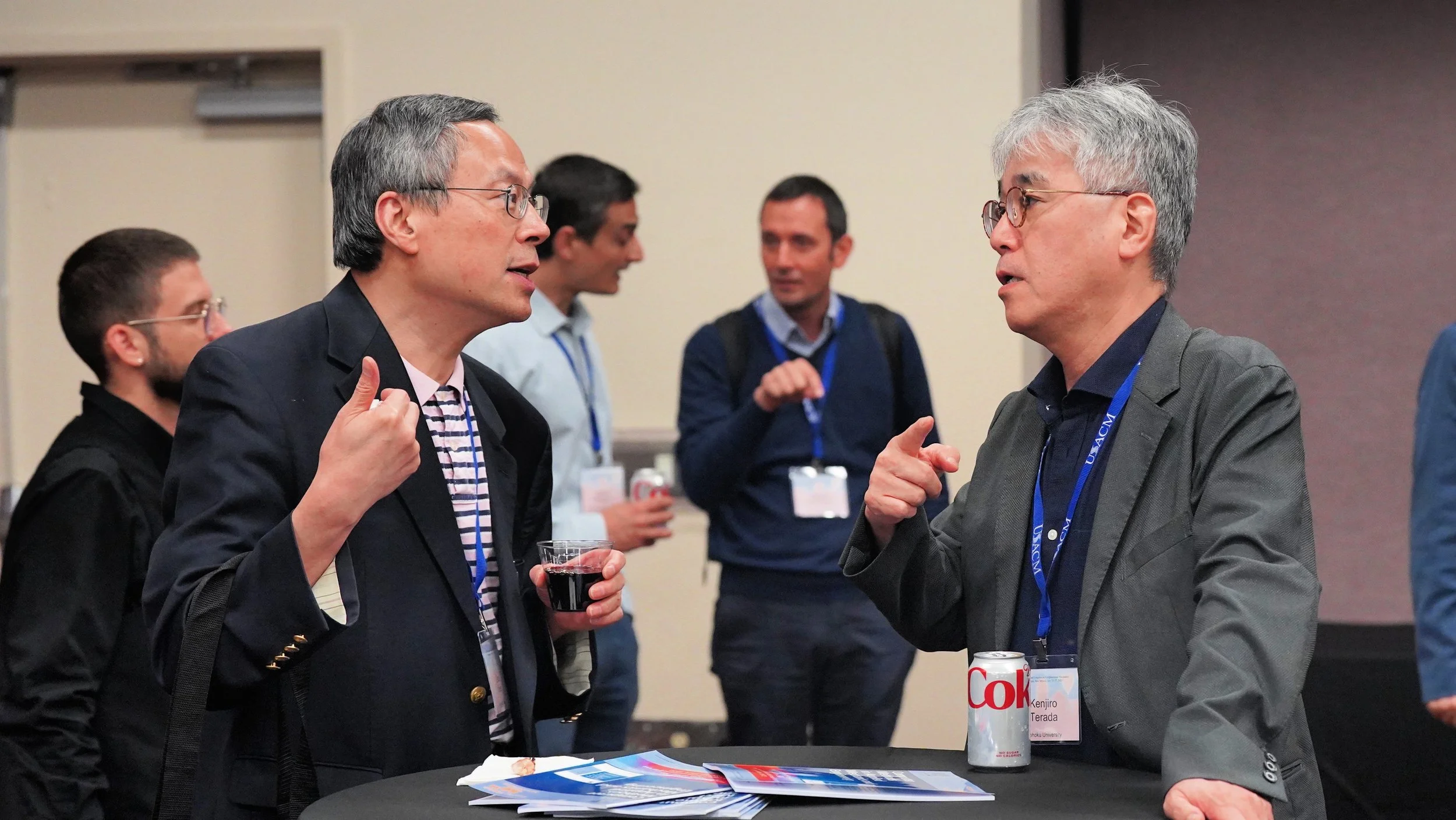 Two men are engaging in a conversation at a conference, with other attendees in the background. They are standing at a round table with pamphlets and a soda can on it.