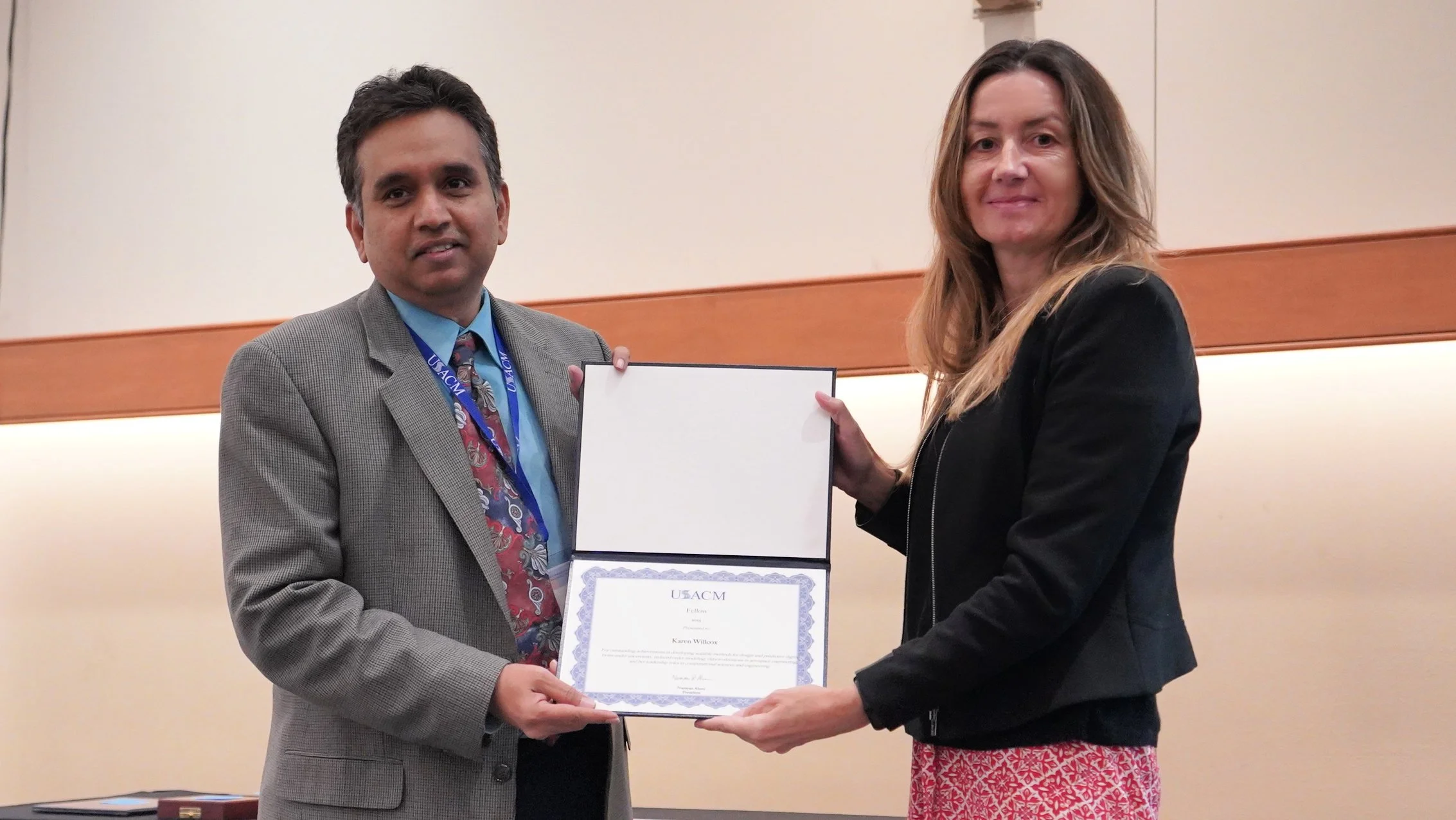 Two people, a man and a woman, standing indoors at an awards ceremony. They are holding a certificate together, with the man on the left and the woman on the right. The man is wearing a grey suit, a blue shirt, a colorful tie, and a blue lanyard. The woman has long wavy hair and is dressed in a black jacket and a patterned red and white skirt. The background features a beige wall with horizontal wooden trim.