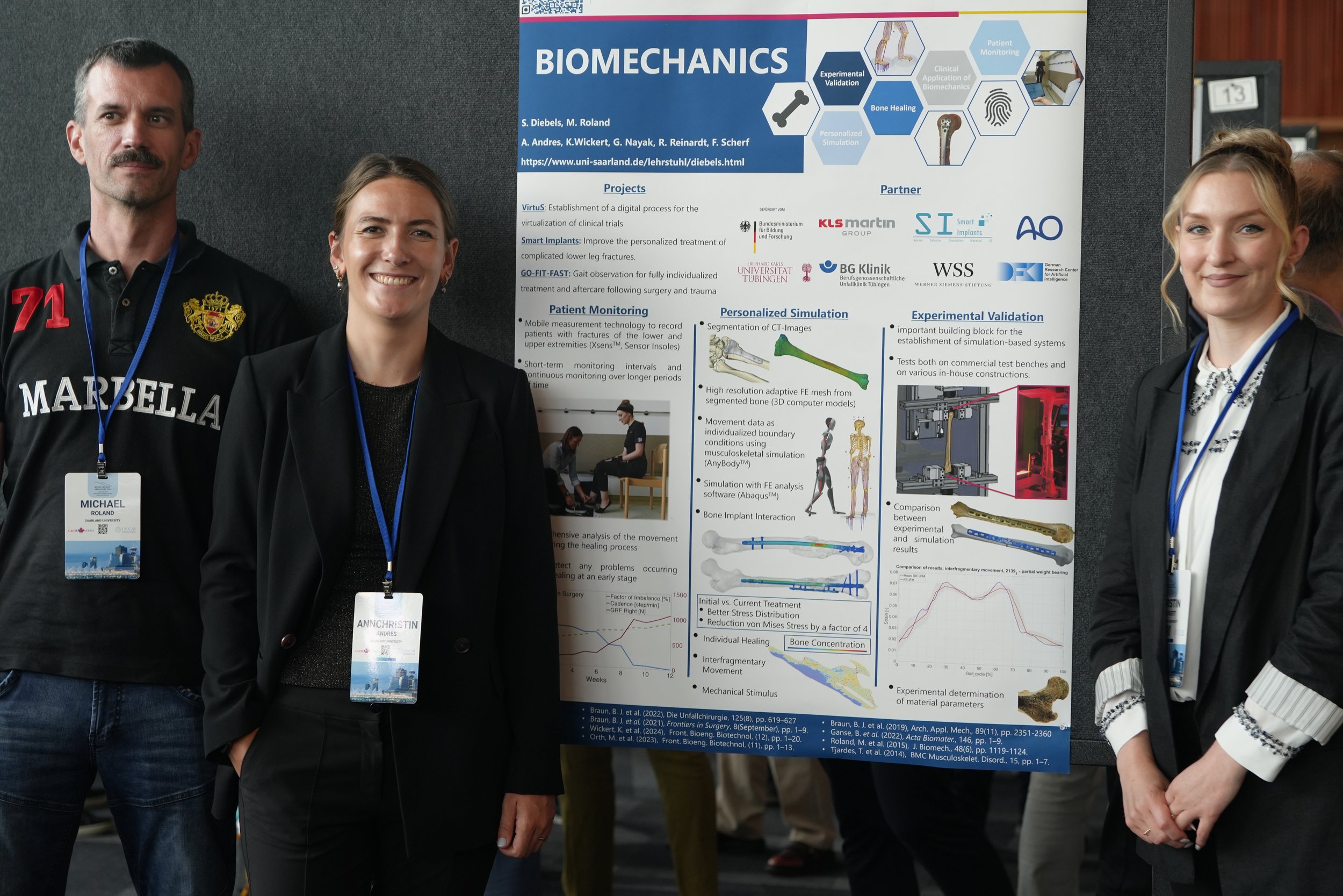 Three people standing next to a scientific research poster at a conference, smiling at the camera. The poster is titled 'Biomechanics' and contains diagrams, charts, and information about research projects related to clinical trials, implants, patient monitoring, and simulation.