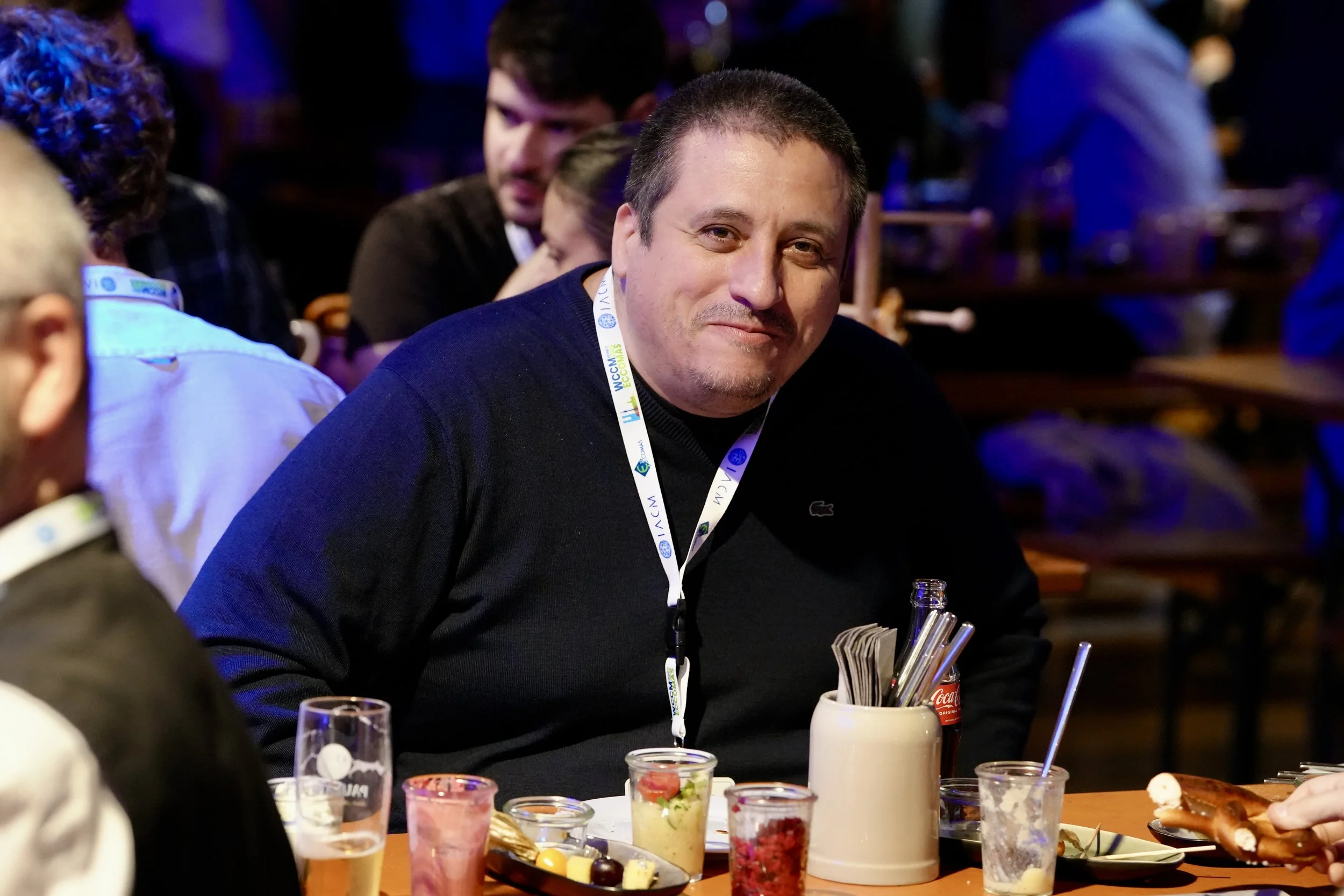 Man smiling at a social gathering seated at a table with various food and drinks, wearing a black shirt with a white lanyard around his neck.
