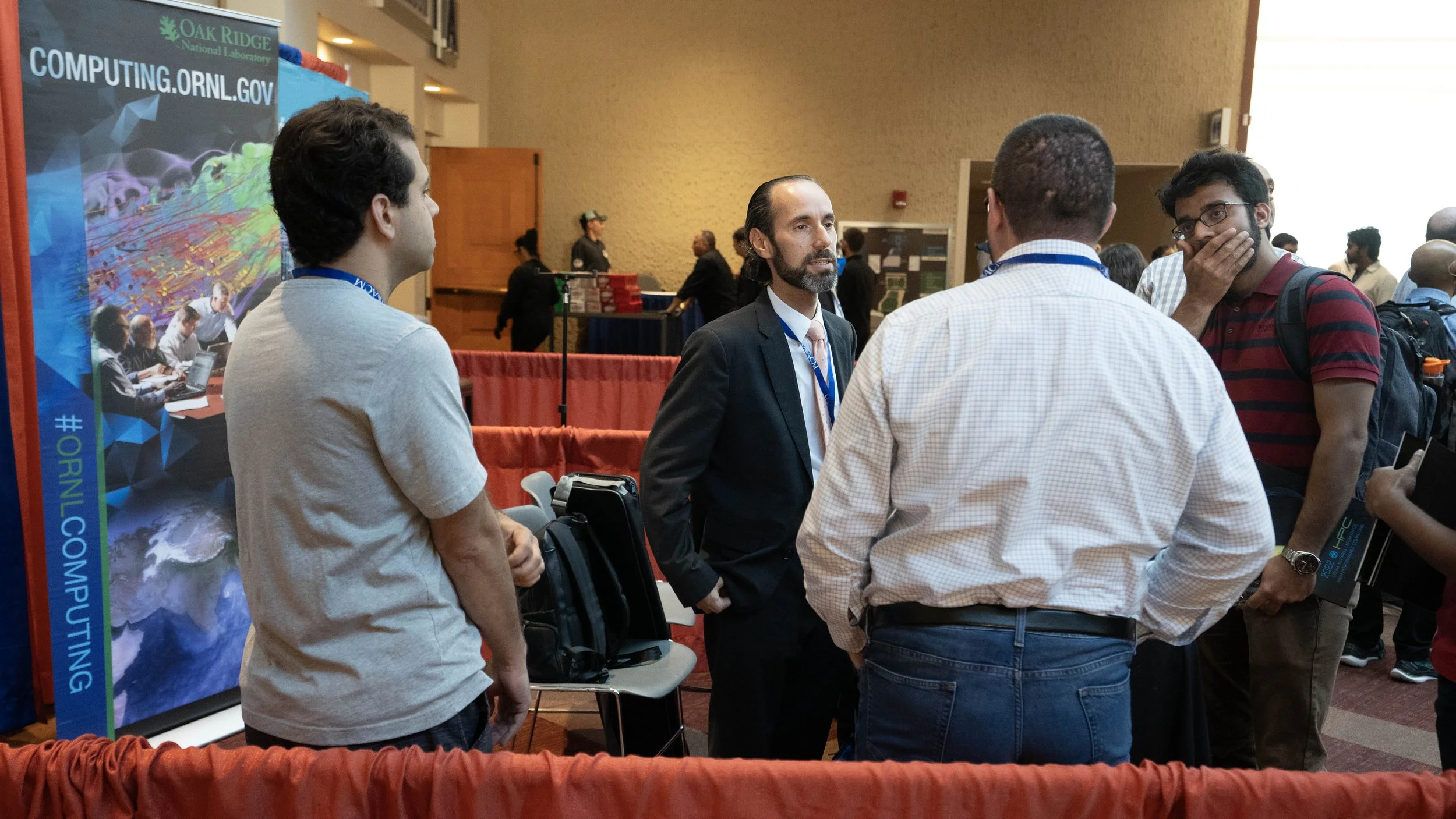 Group of four men engaging in conversation at a conference, with one man in a suit and three casually dressed, near a banner for Oak Ridge National Laboratory.