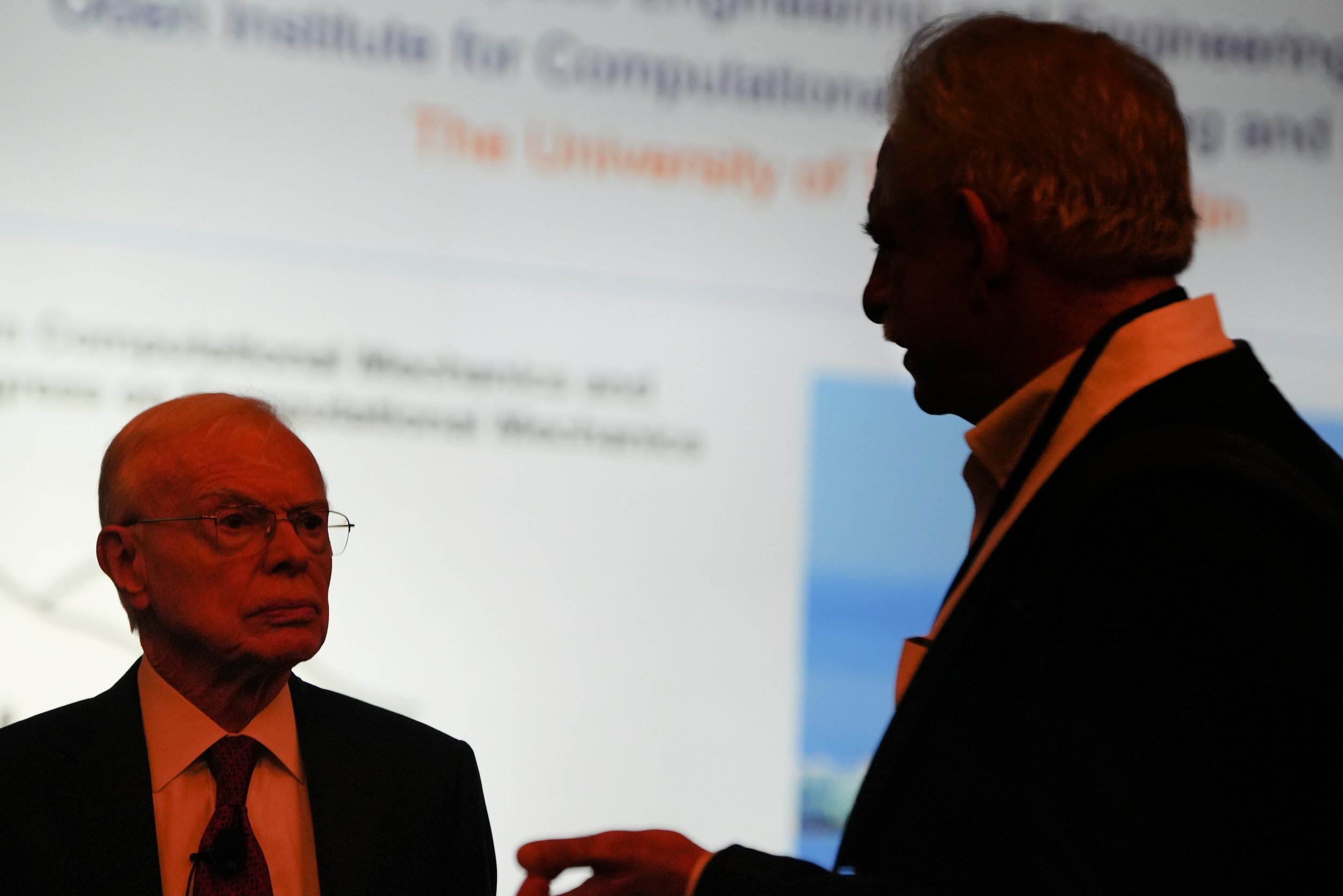 Two men in a dark room with a projected presentation in the background. One man is older, wearing glasses and a suit with a t-shirt underneath, and looking at the other man. The second man is younger, with light-colored hair, dressed in a suit with a collared shirt, and appears to be speaking or explaining something.