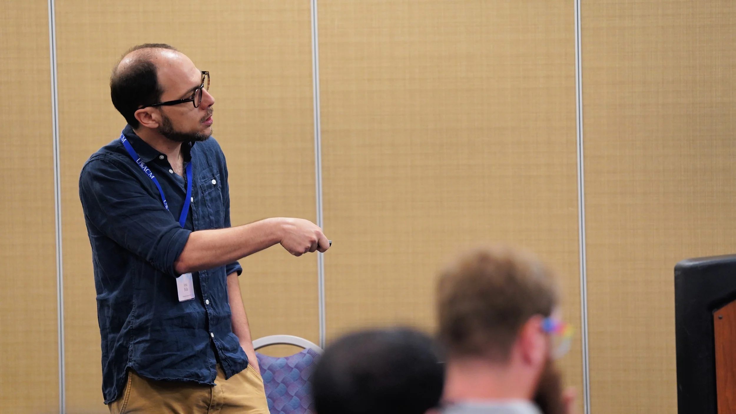 A man with glasses and a beard standing in front of a yellow partition, giving a presentation to an audience in a conference room.