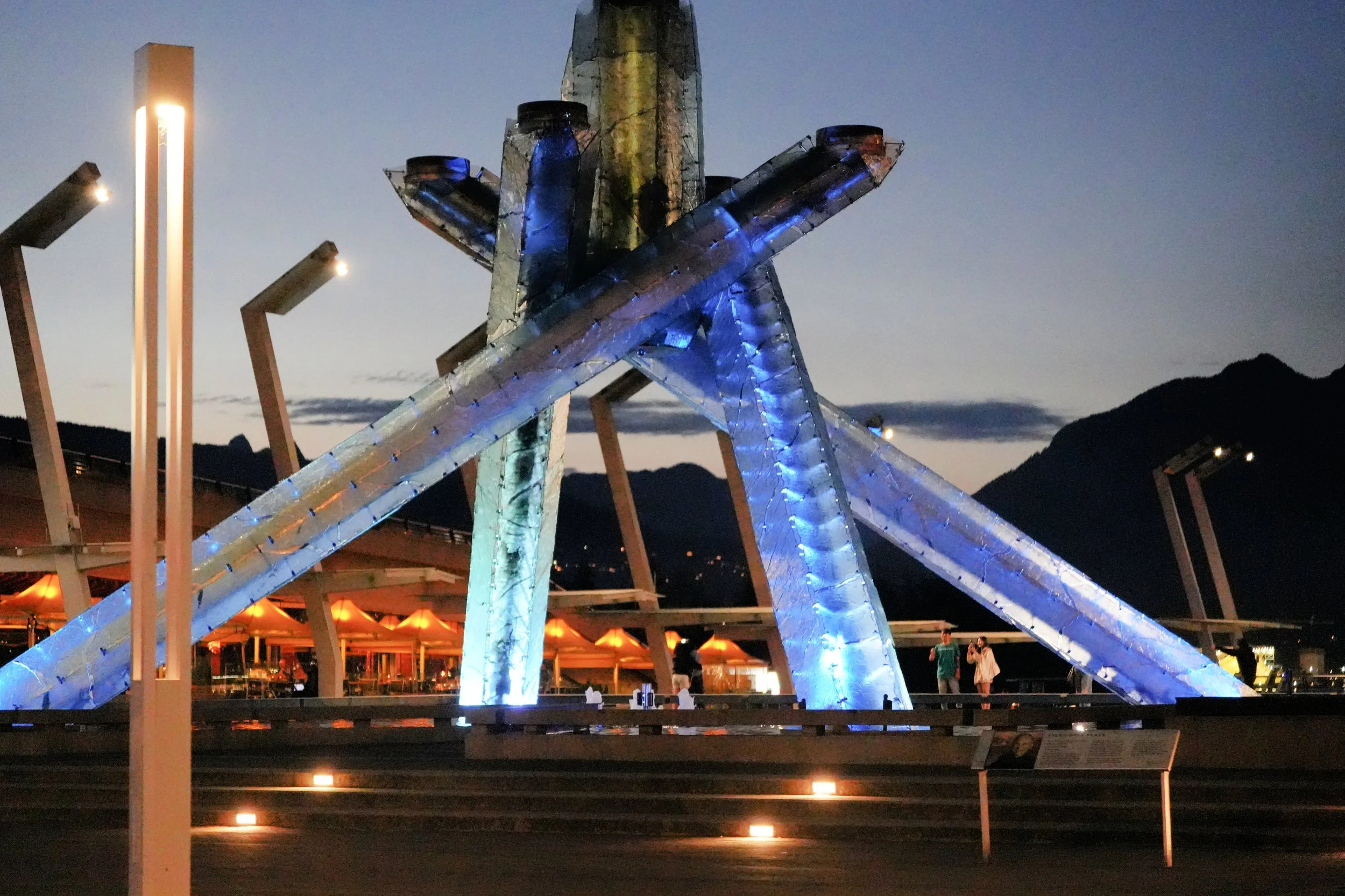 Nighttime photo of a star-shaped amusement park ride illuminated with blue and white lights, with mountains in the background and a few people nearby.