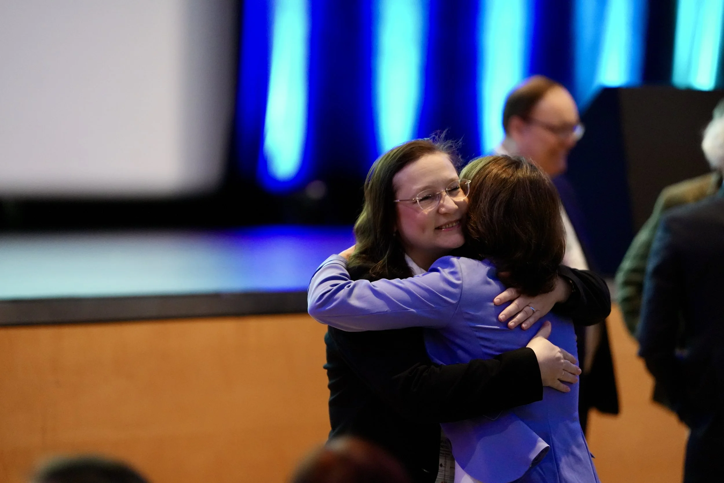 Two women hugging each other warmly, smiling, during a session at an academic conference.
