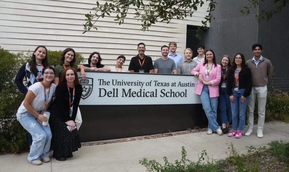 Group of people standing behind a sign that reads "The University of Texas at Austin Dell Medical School," outdoors near a building and greenery.