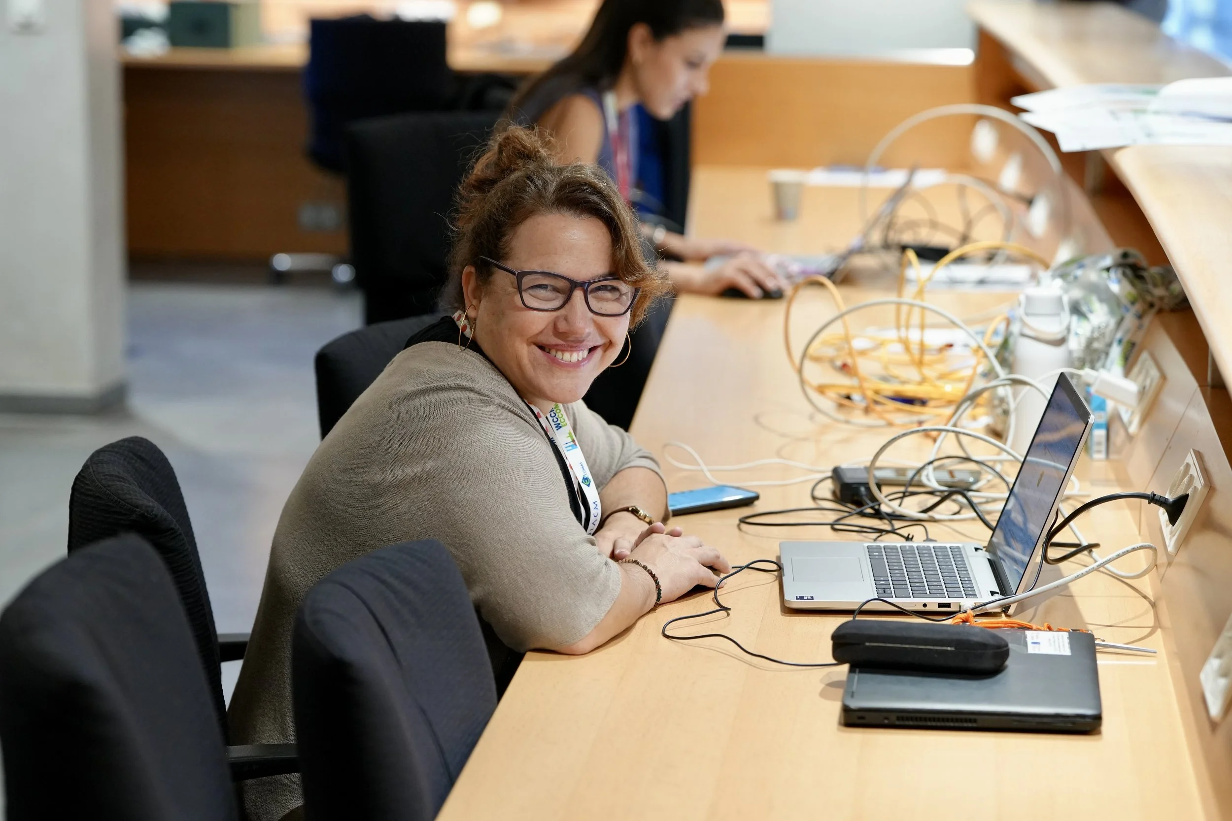 Woman with glasses smiling at a desk with laptops and wires in an office setting.