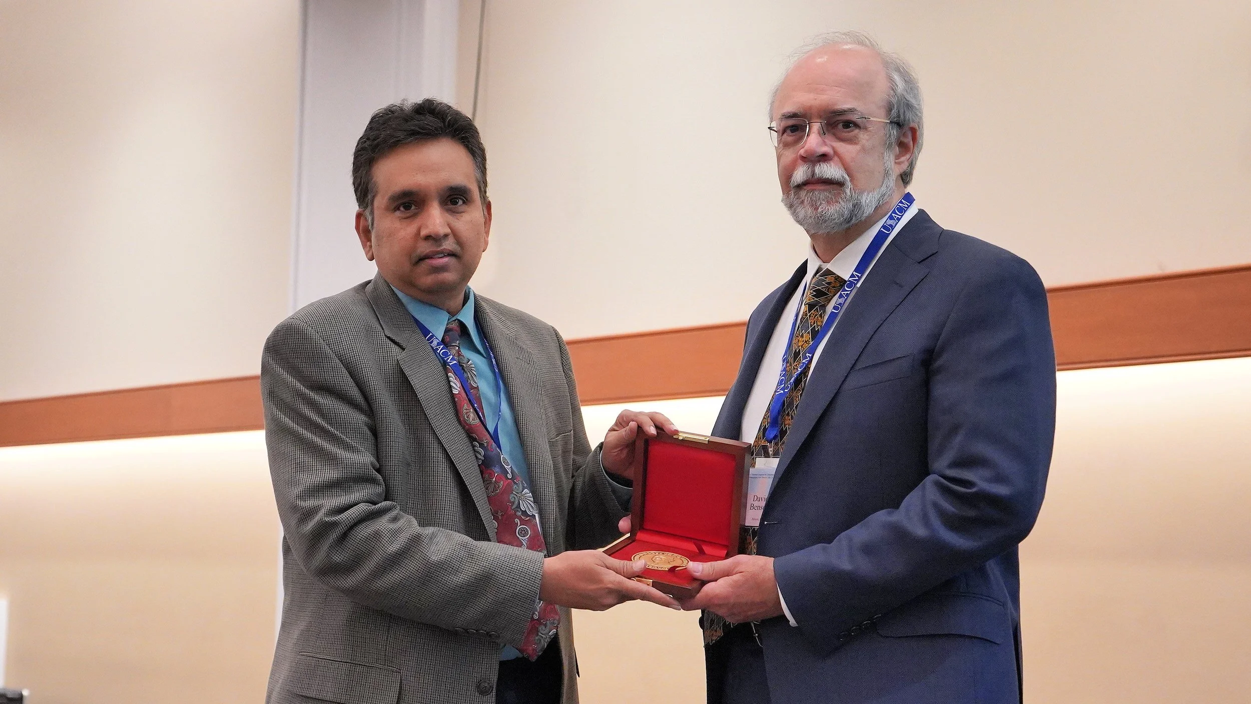 Two men in suits shaking hands, one is presenting an award or medal in a red box to the other inside a room with beige walls.