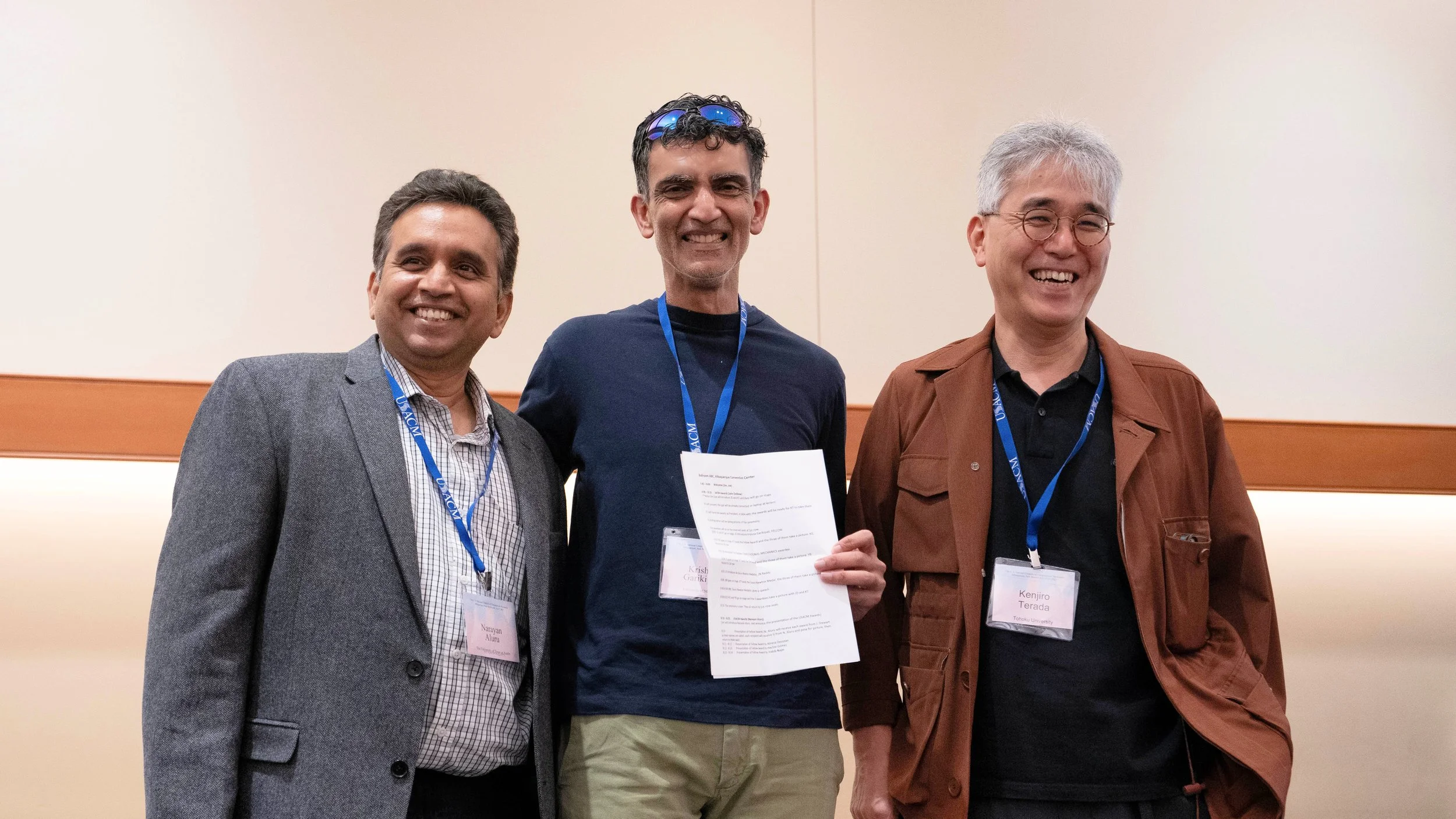Three men standing next to each other at an indoor event, smiling. They are wearing conference badges on lanyards. The man on the left is in a gray suit jacket with a plaid shirt, the man in the middle is in a navy blue shirt holding a paper, and the man on the right is in a brown jacket with glasses.