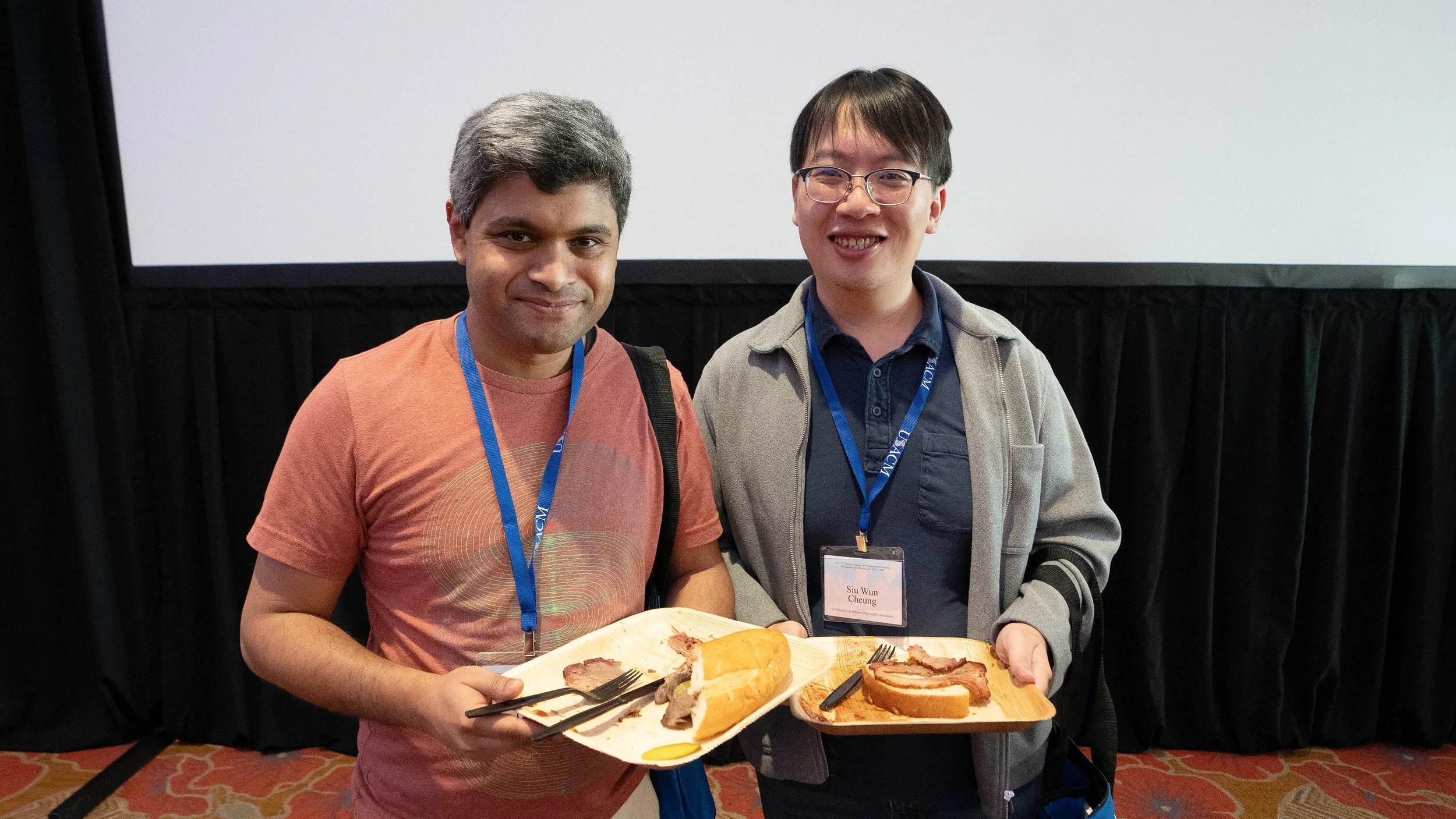 Two men standing together holding a plate of food including slices of roast beef and bread, smiling at the camera, in an indoor setting with a white screen and black curtains behind them.