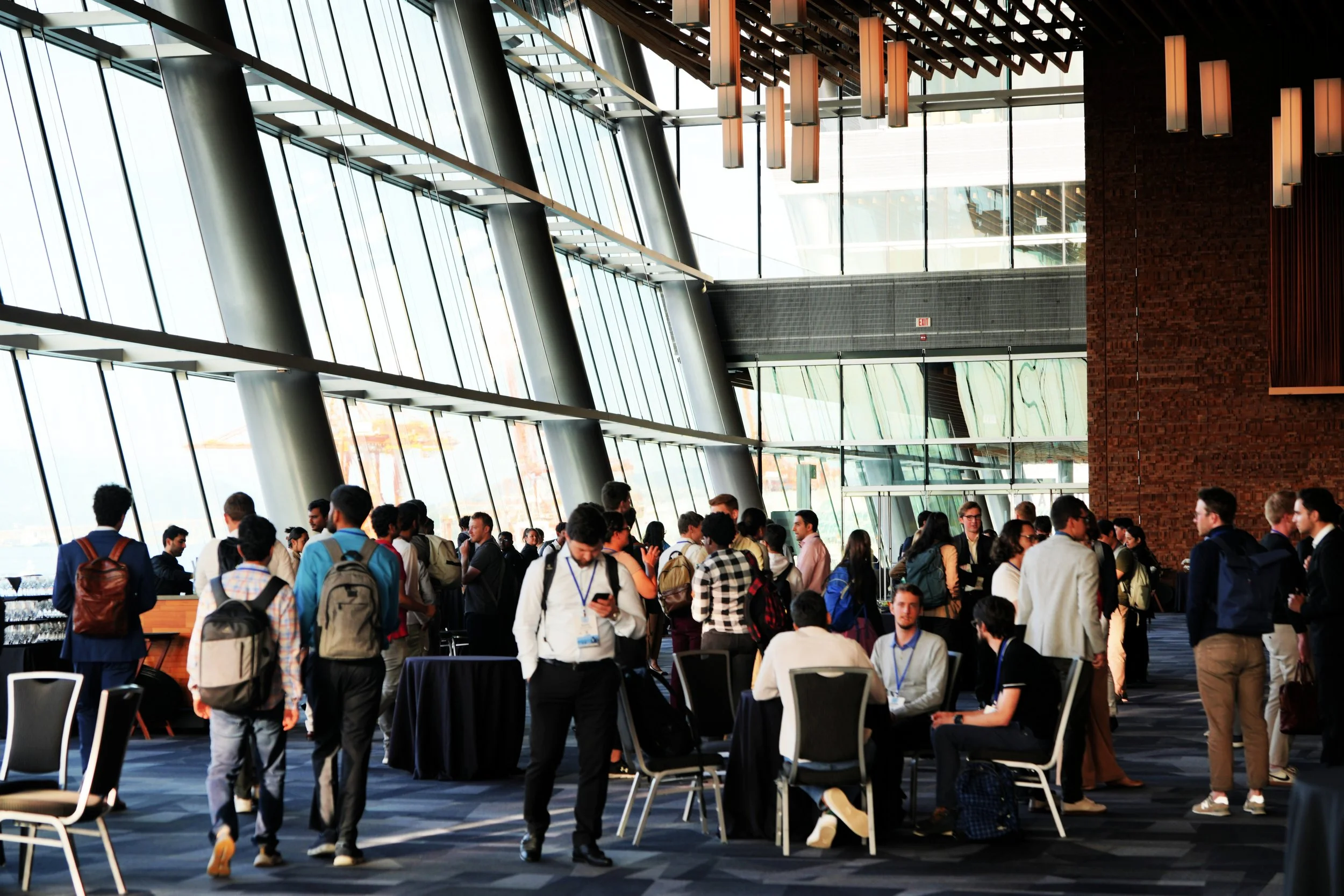 A large indoor conference or convention center filled with people, with high glass ceilings and modern architecture, some standing and some sitting, engaged in conversations or using devices.