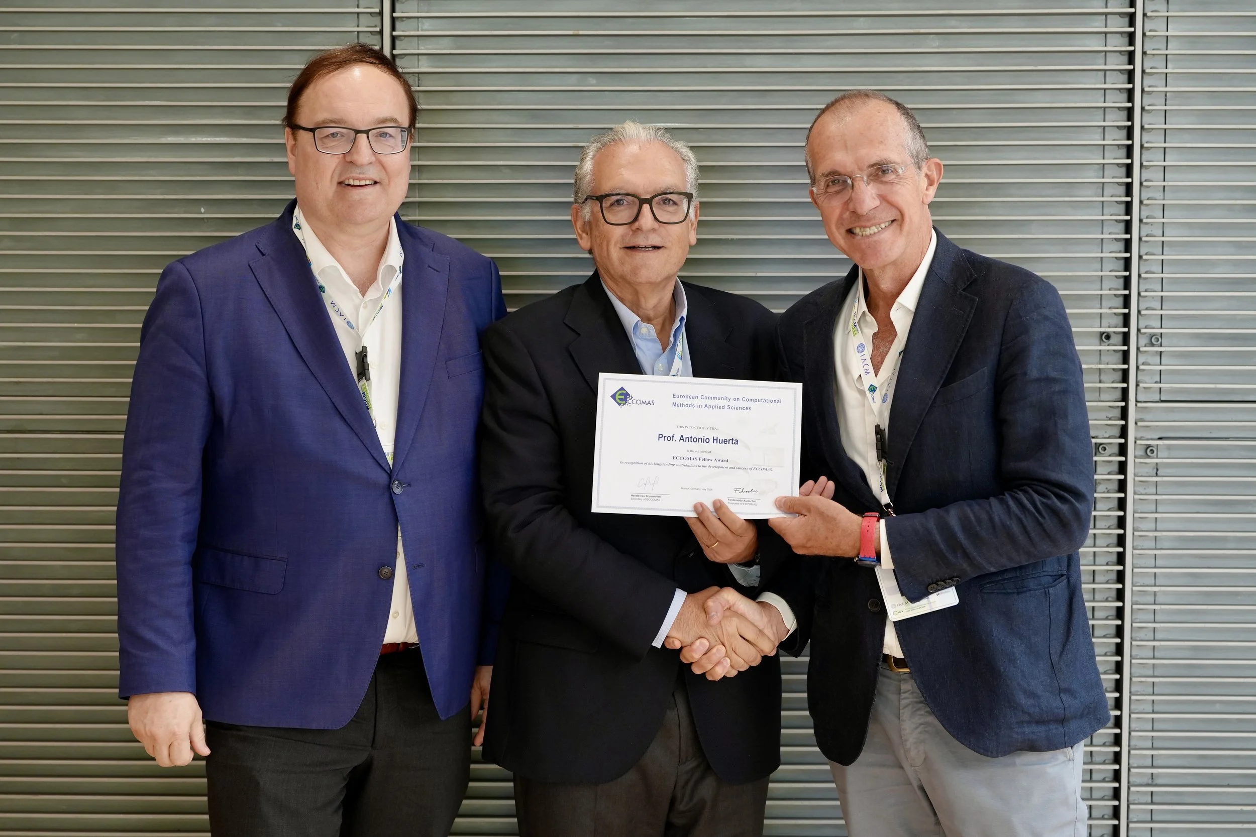 Three men in suits at a conference, one holding a certificate. They are standing in front of a metal slatted wall, smiling and shaking hands.