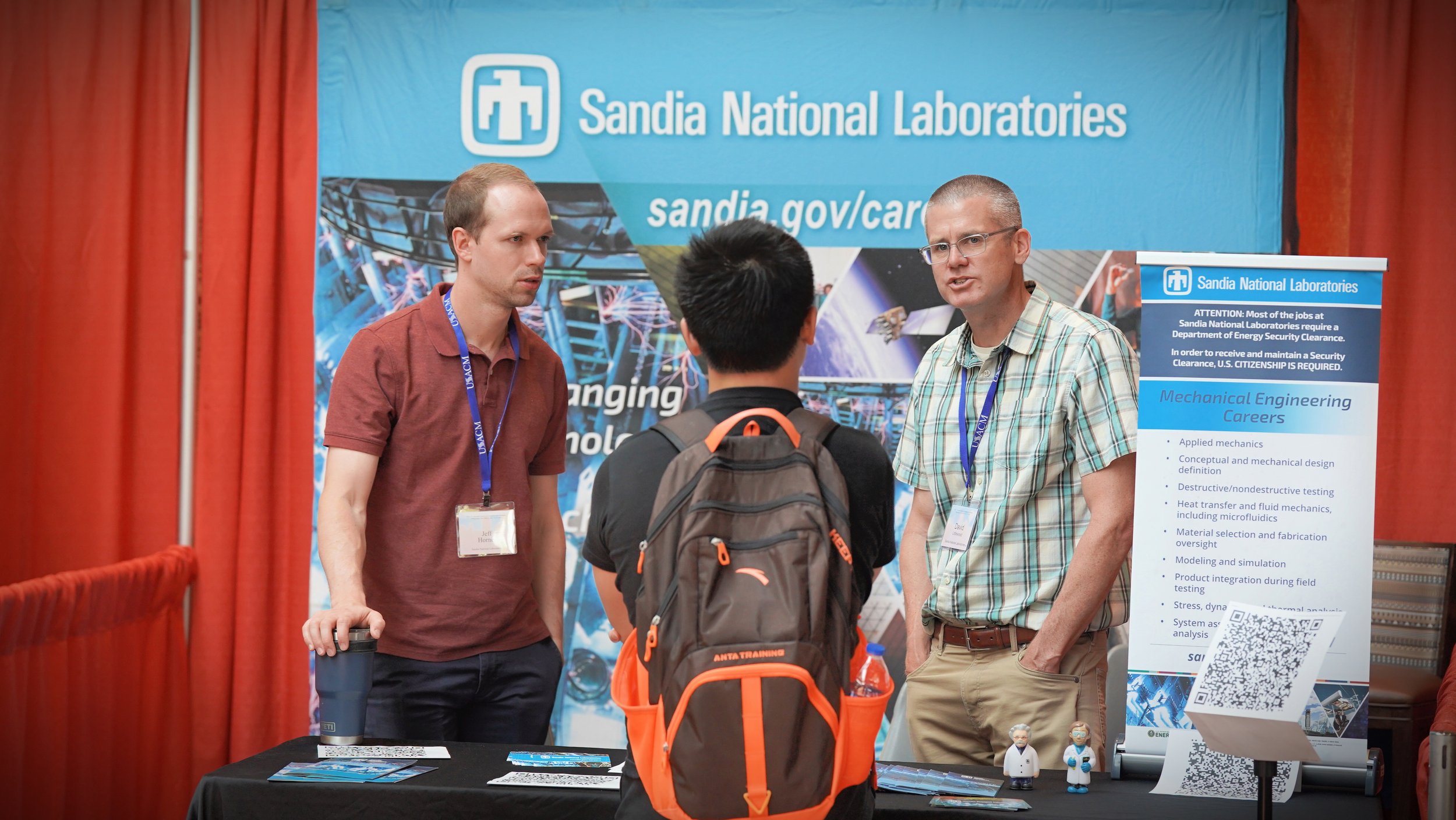 A group of men engaged in conversation at a Sandia National Laboratories booth at an event. The booth has a large blue banner displaying the Sandia logo and information about careers in mechanical engineering, with red curtains in the background.