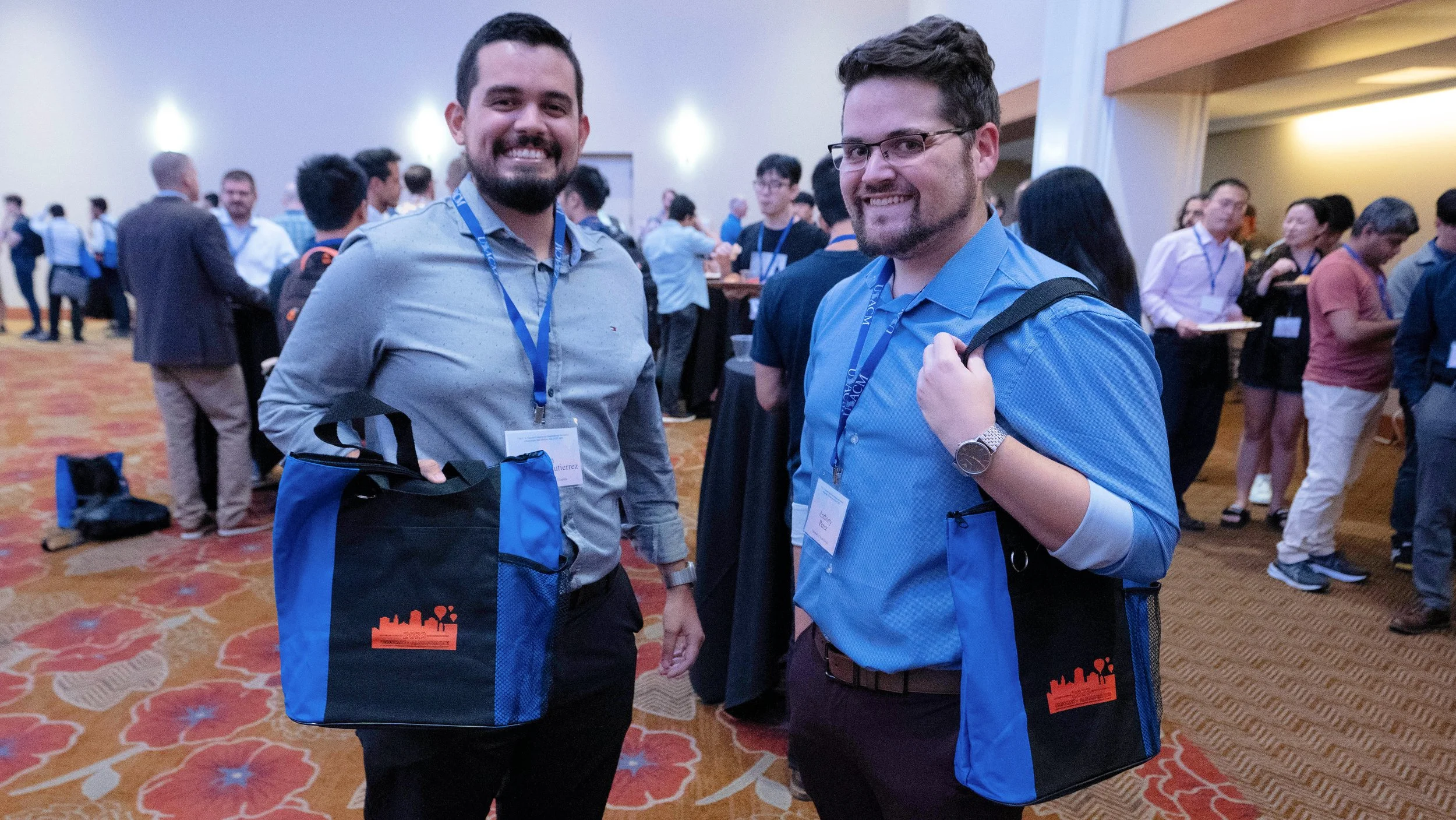 Two men smiling at a conference, carrying bags, with many people in the background.