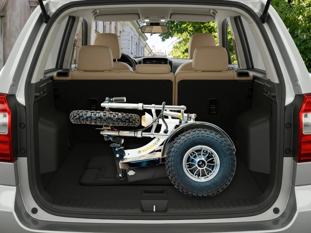 The rear view of a vehicle showing an off-road wheelchair with large tires and a white frame, positioned inside the trunk, with beige seats and a navigation system visible through the windshield.