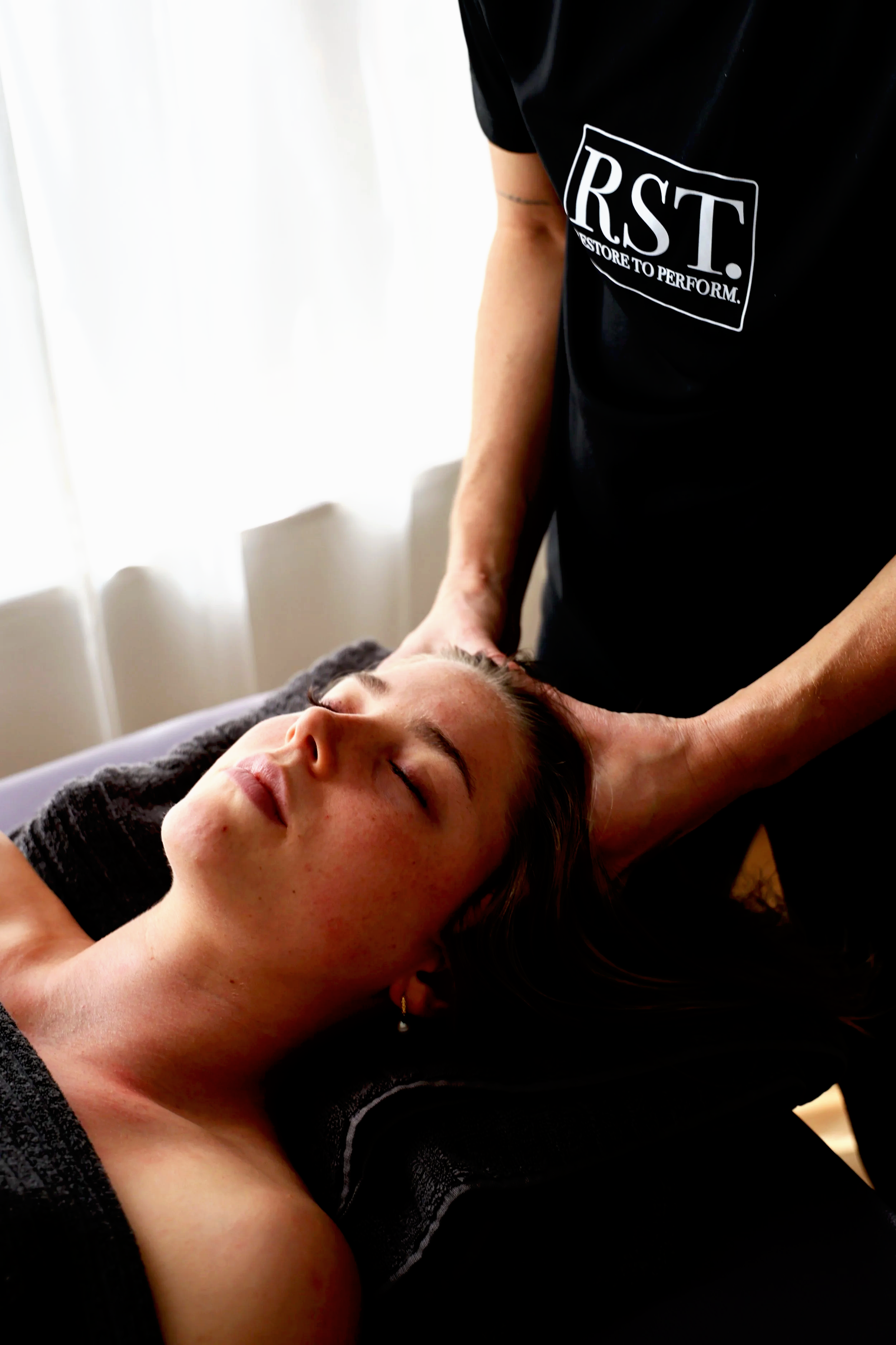 A woman lying on her back with eyes closed, receiving a massage or therapy from a practitioner wearing a black shirt with the text 'RST. Restore to Perform.' on it, in a bright room with white curtains.