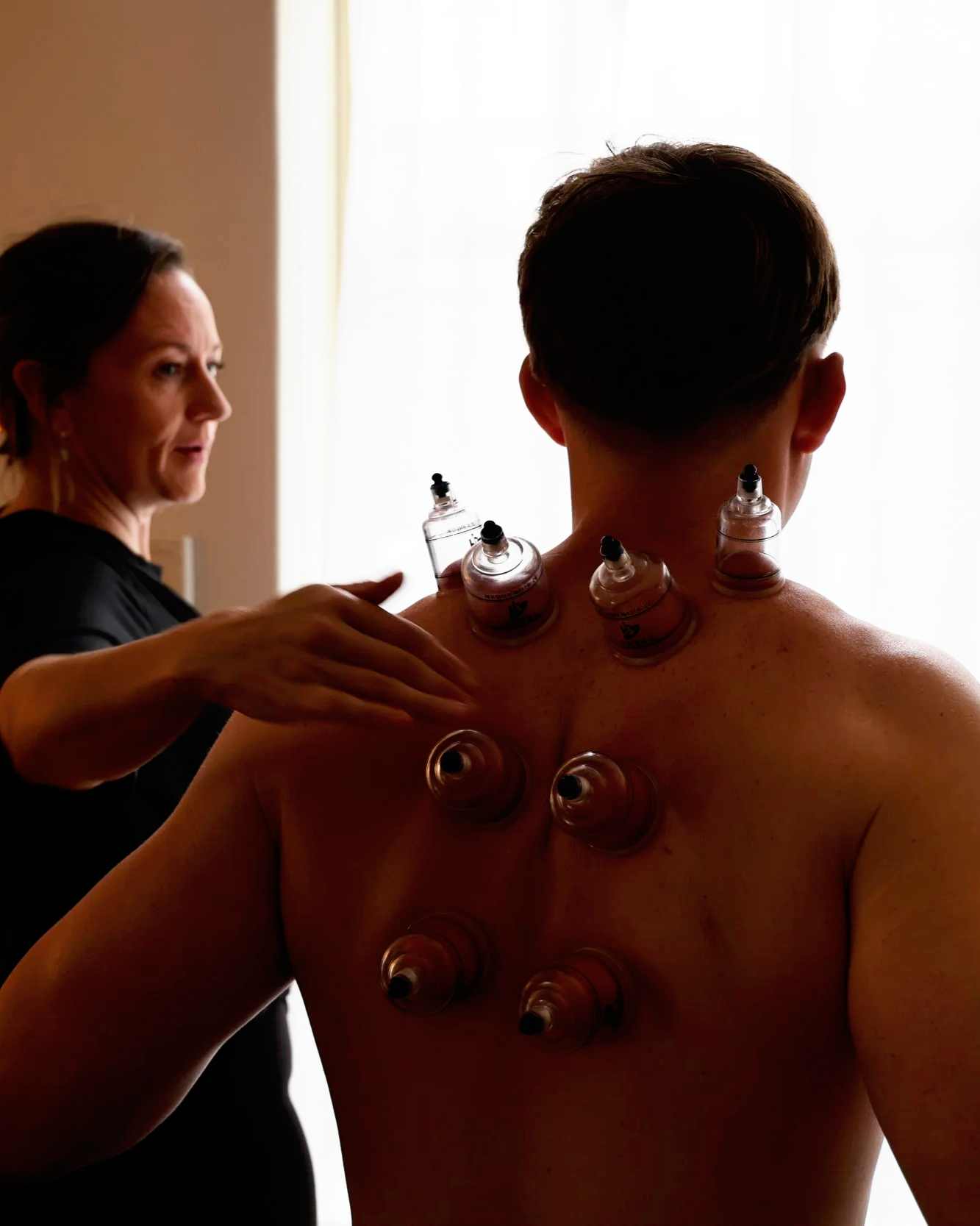 Woman with short brown hair having plastic cups placed on her back by a therapist, in front of a window with bright light.