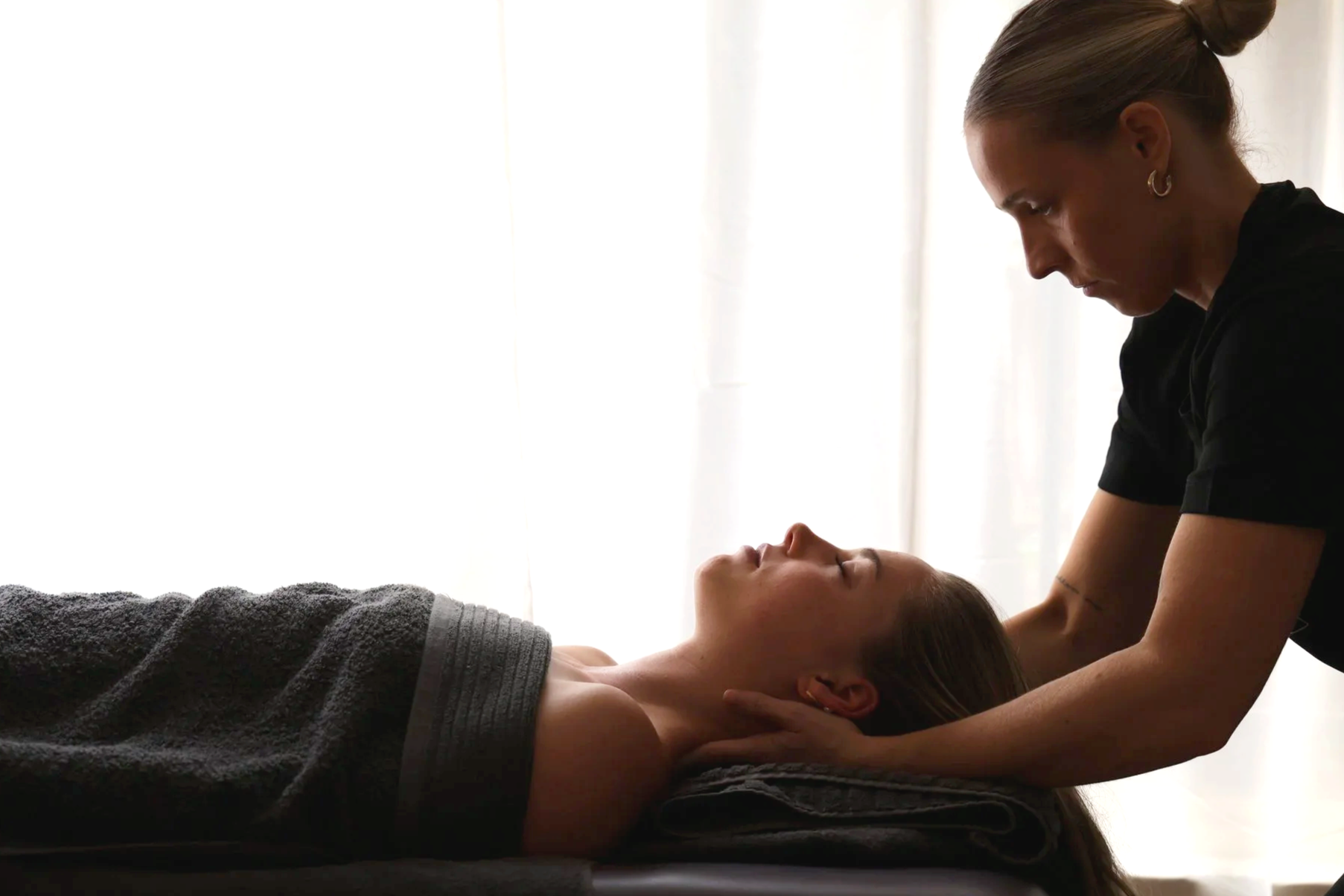 A woman lying on her back on a massage table with her eyes closed, receiving a massage from a woman leaning over her, in front of a bright window with white curtains.