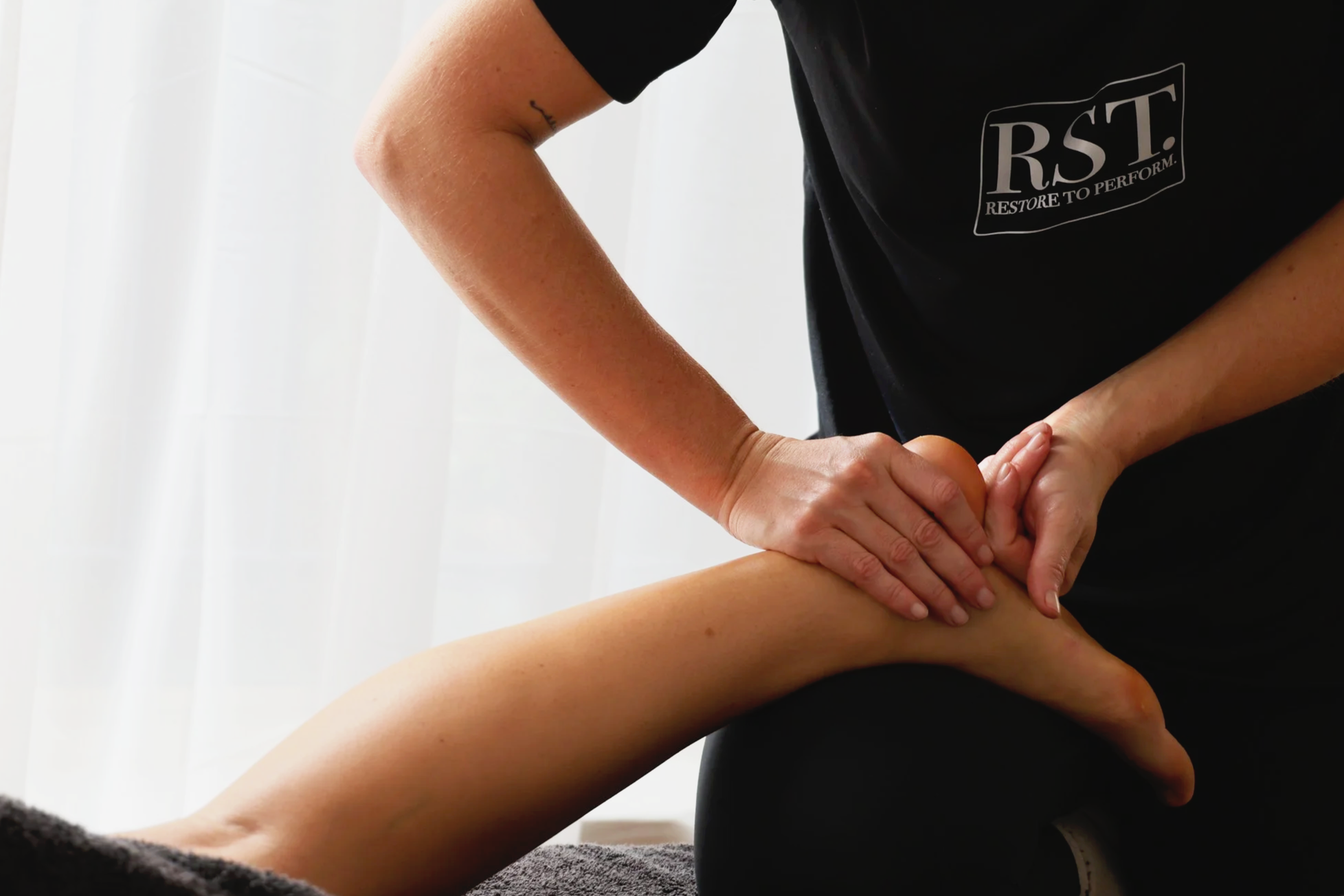 A massage therapist performing a massage on a client's leg in a well-lit room with white curtains. The therapist wears a black shirt with a logo that reads 'RST.' The client's leg is being massaged with hands applying pressure, and the client appears relaxed.
