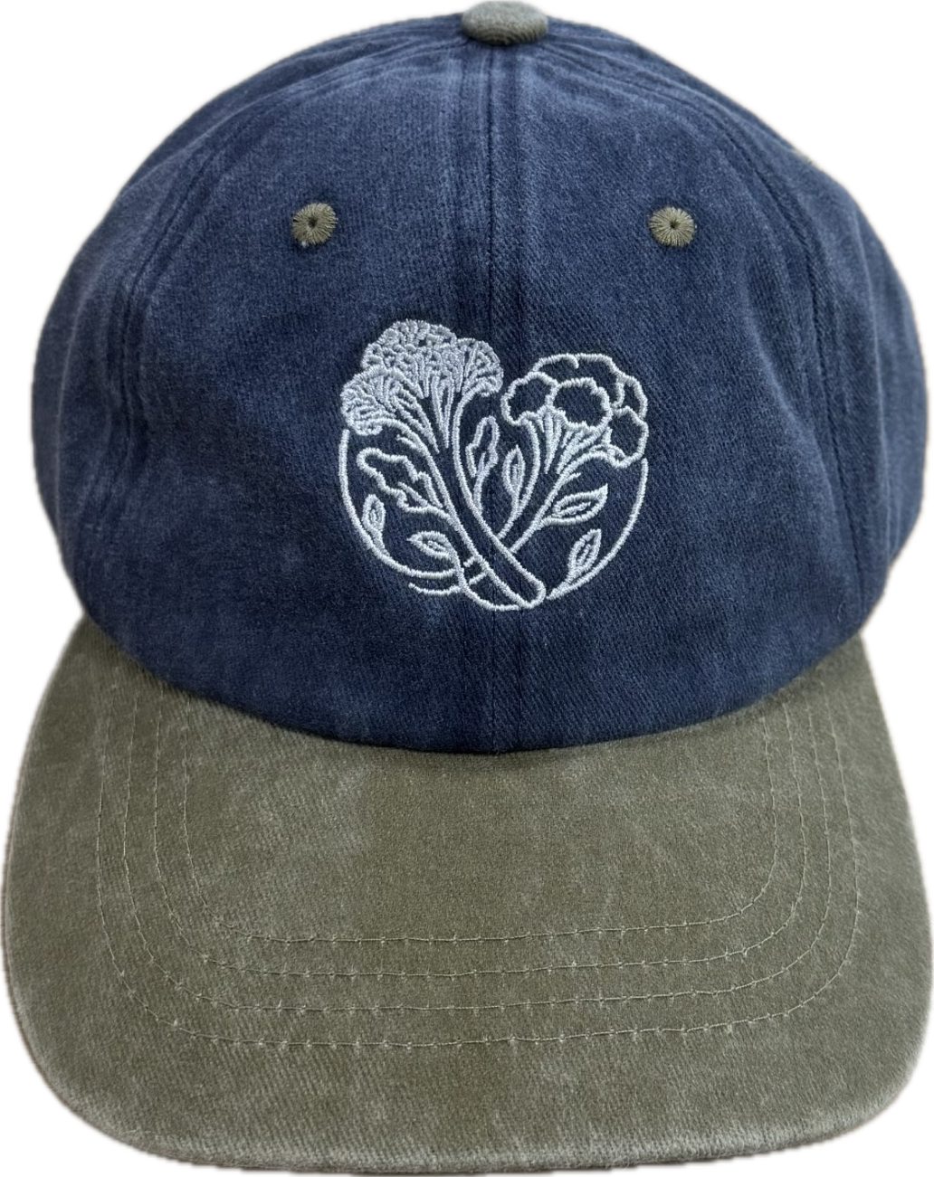 A baseball cap with a blue crown, a light grey brim, and embroidered white floral design on the front.