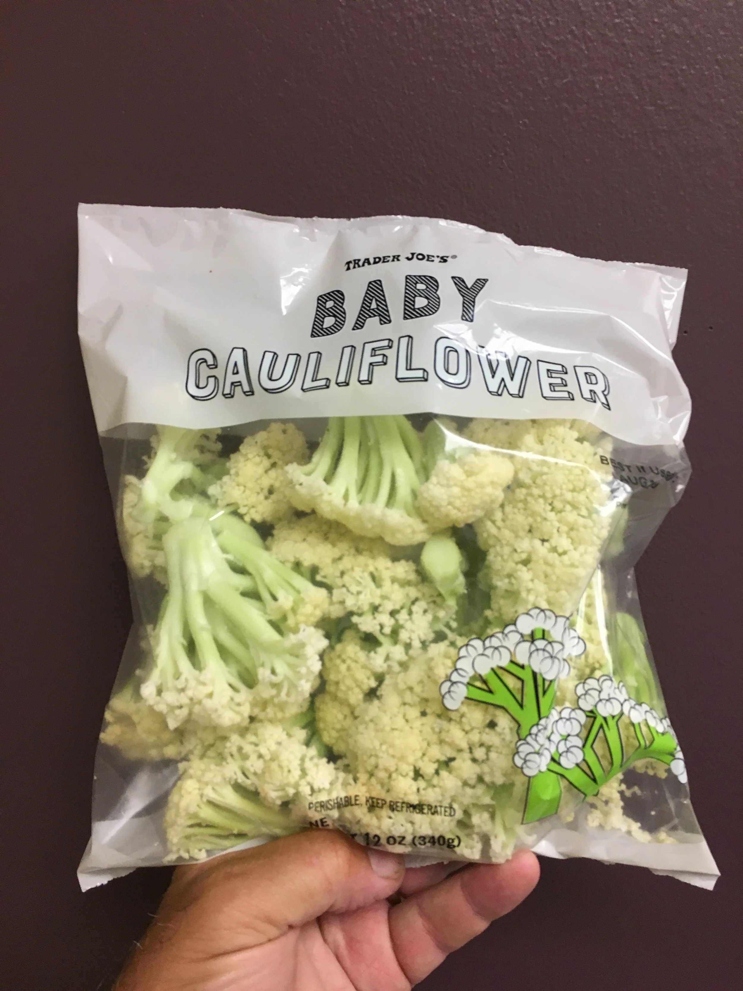 A hand holding a bag of Trader Joe's baby cauliflower against a dark background.