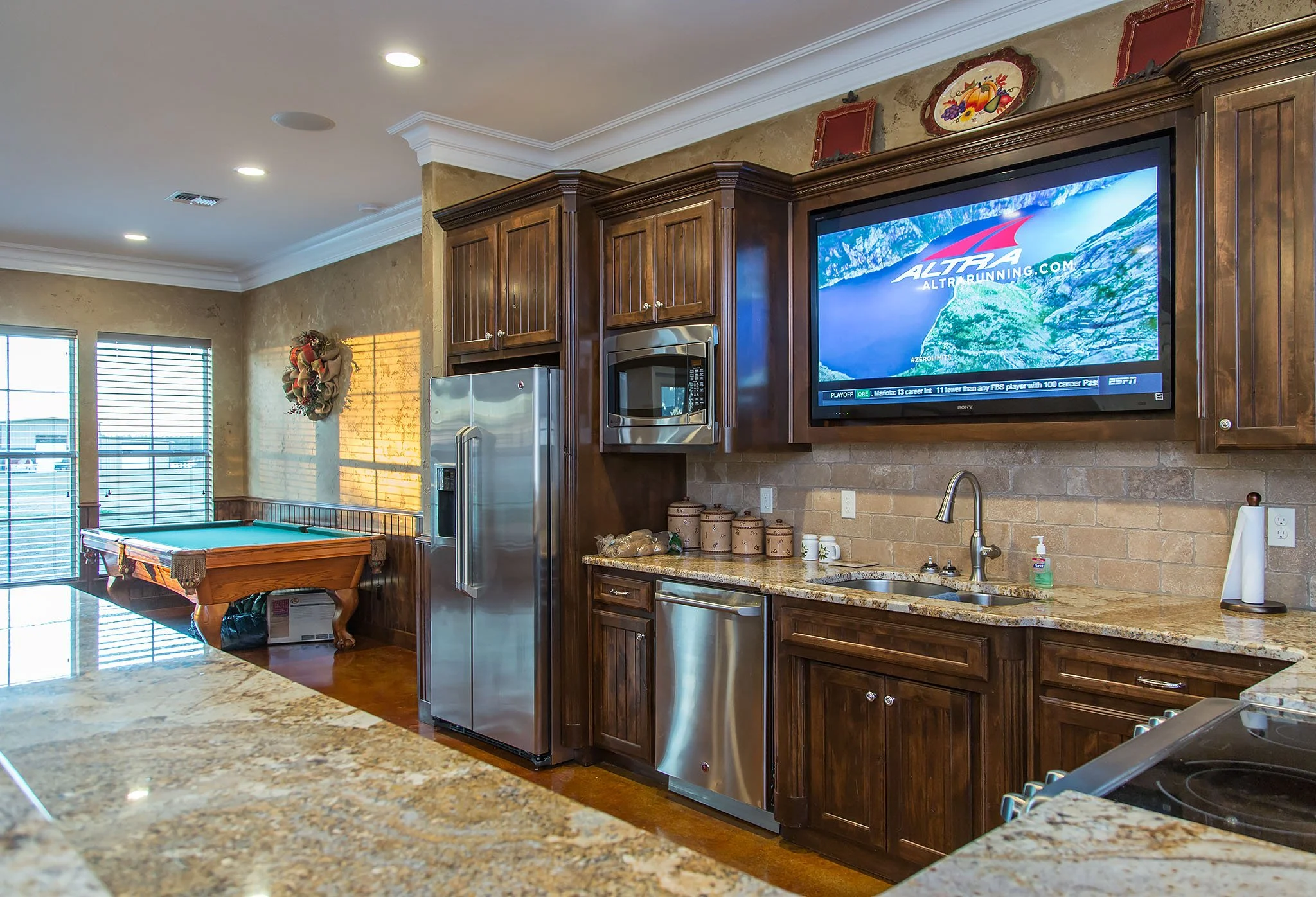 A kitchen with wooden cabinets, a granite countertop, a stainless steel refrigerator, a microwave oven, a sink with a faucet, a TV on the wall displaying a sports channel, and a pool table in the room. There are colorful plates and decorative items above the cabinets and a wreath on the wall near a sliding door with blinds.